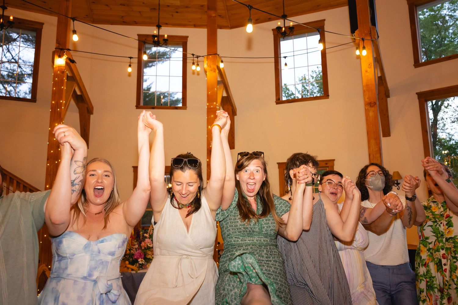Joyful wedding guests raise their arms in celebration during a reception in a rustic indoor venue.
