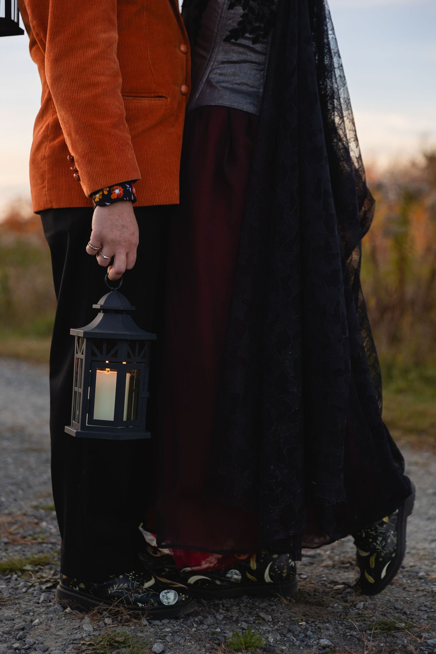 Two people in contrasting orange and black outfits embrace while holding a decorative lantern outdoors.