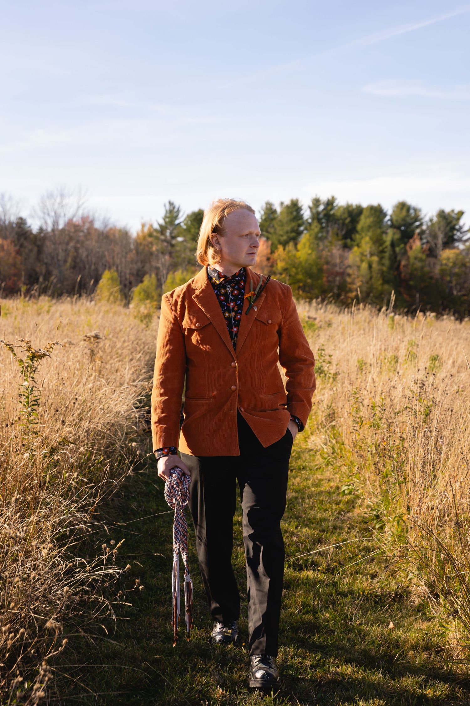 Person in brown jacket walking through tall autumn grass field at golden hour with trees in background.