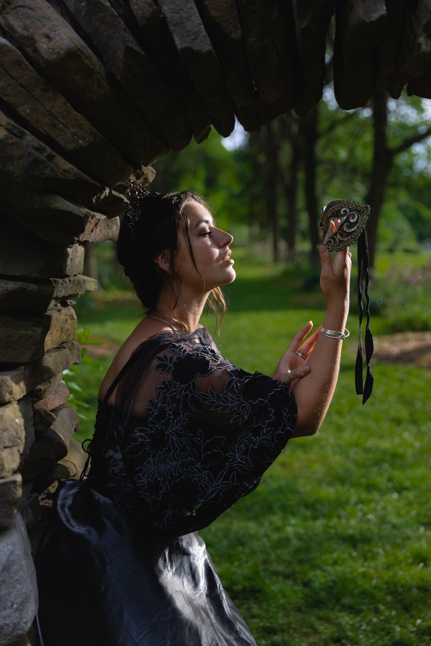 Woman in black lace dress holding ornate mirror beside stone wall outdoors.