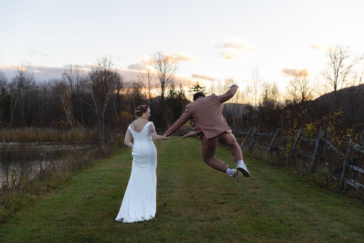 One person jumps joyfully while other holds dress during sunset wedding photo session.