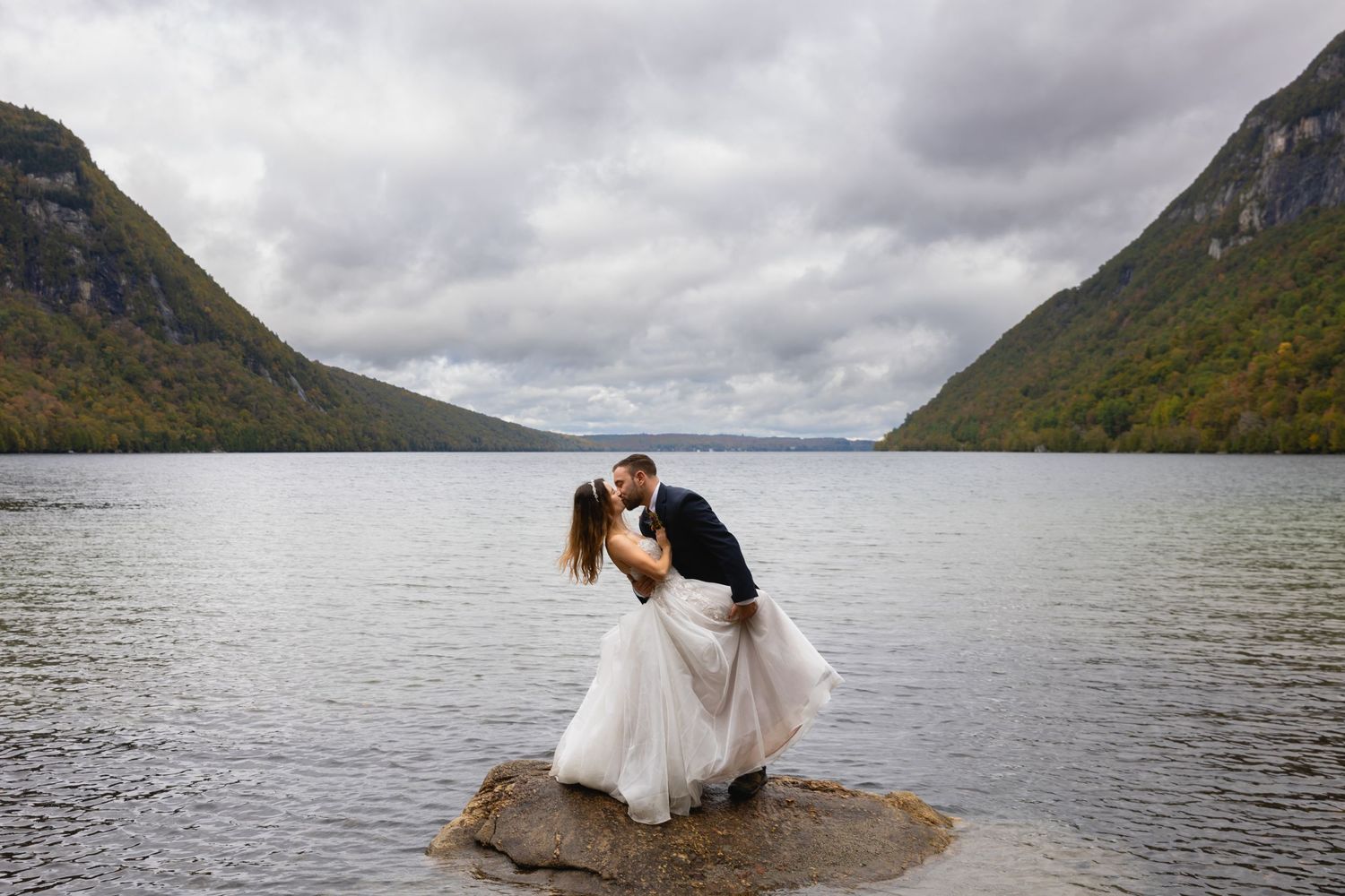 Romantic wedding couple embracing on rock in mountain lake during cloudy day.