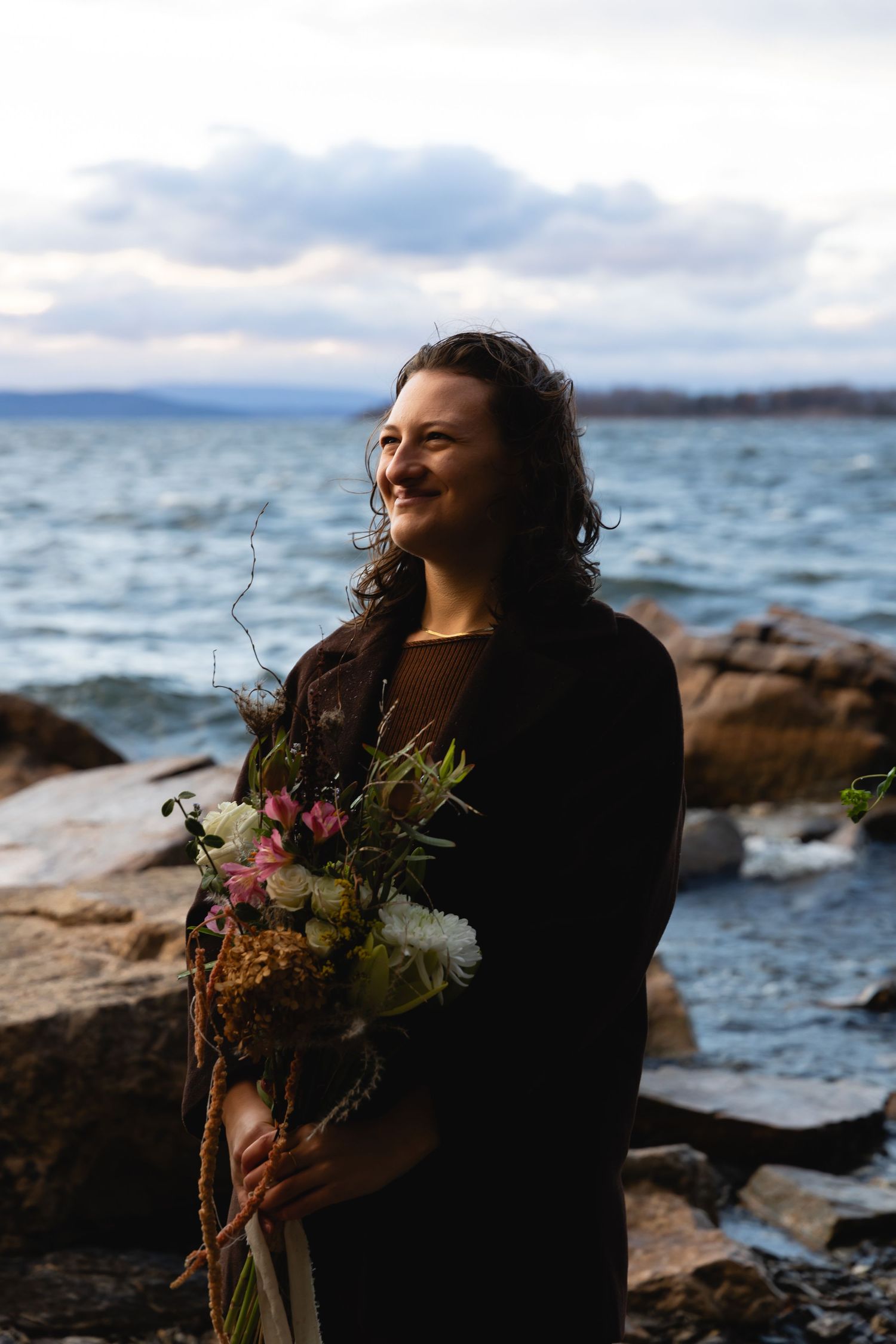 Someone wearing a black coat holds a beautiful wildflower bouquet while standing on rocky coastline at sunset.