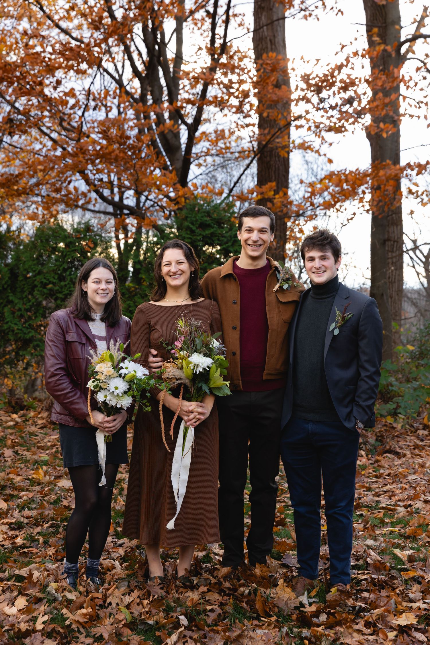 A group of four young people stand among fallen autumn leaves wearing casual attire and holding flower bouquets.