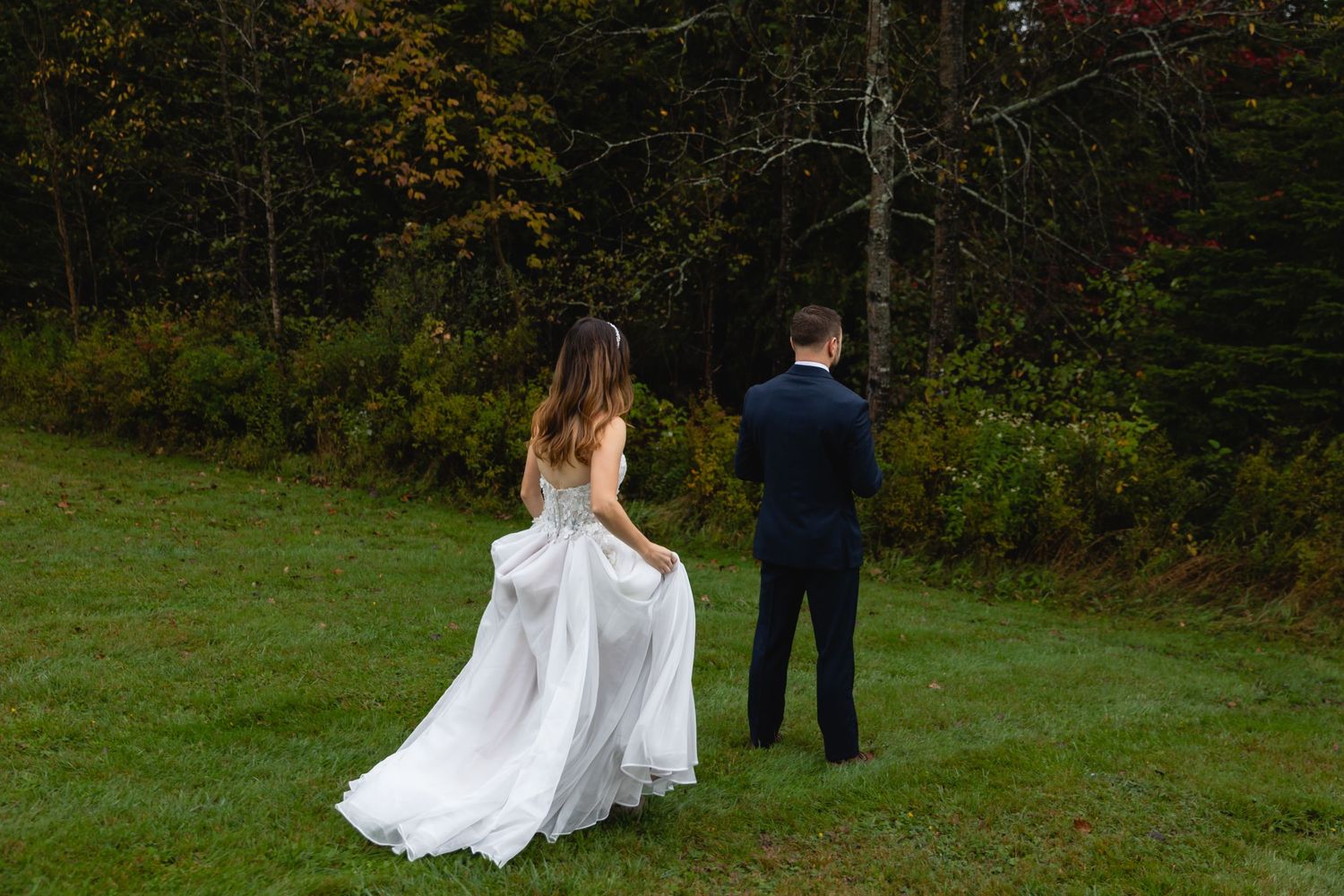 A couple in wedding attire stands facing away in a green autumn field with trees.