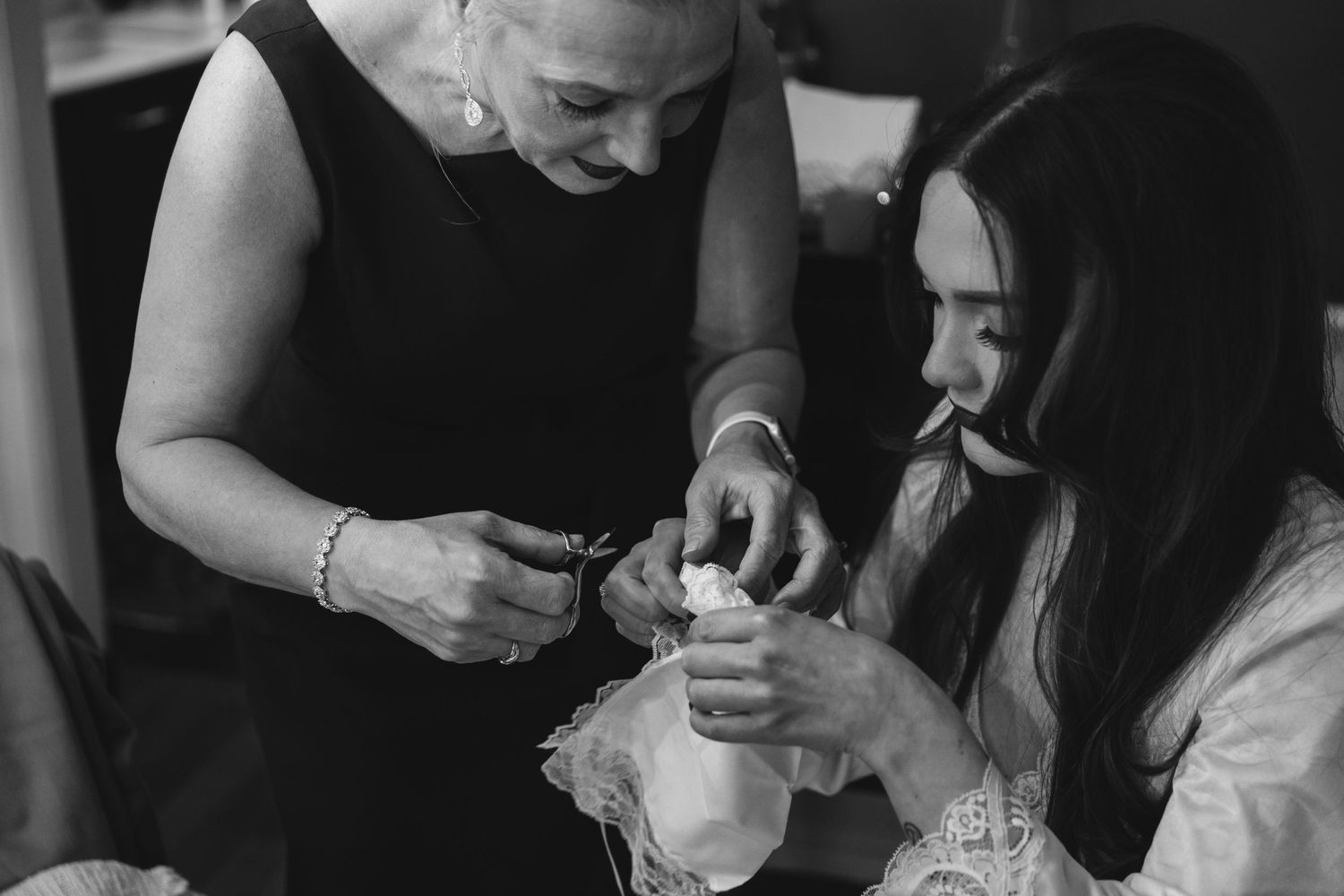 A black and white photograph shows two people working together to fix details on a delicate piece of clothing.
