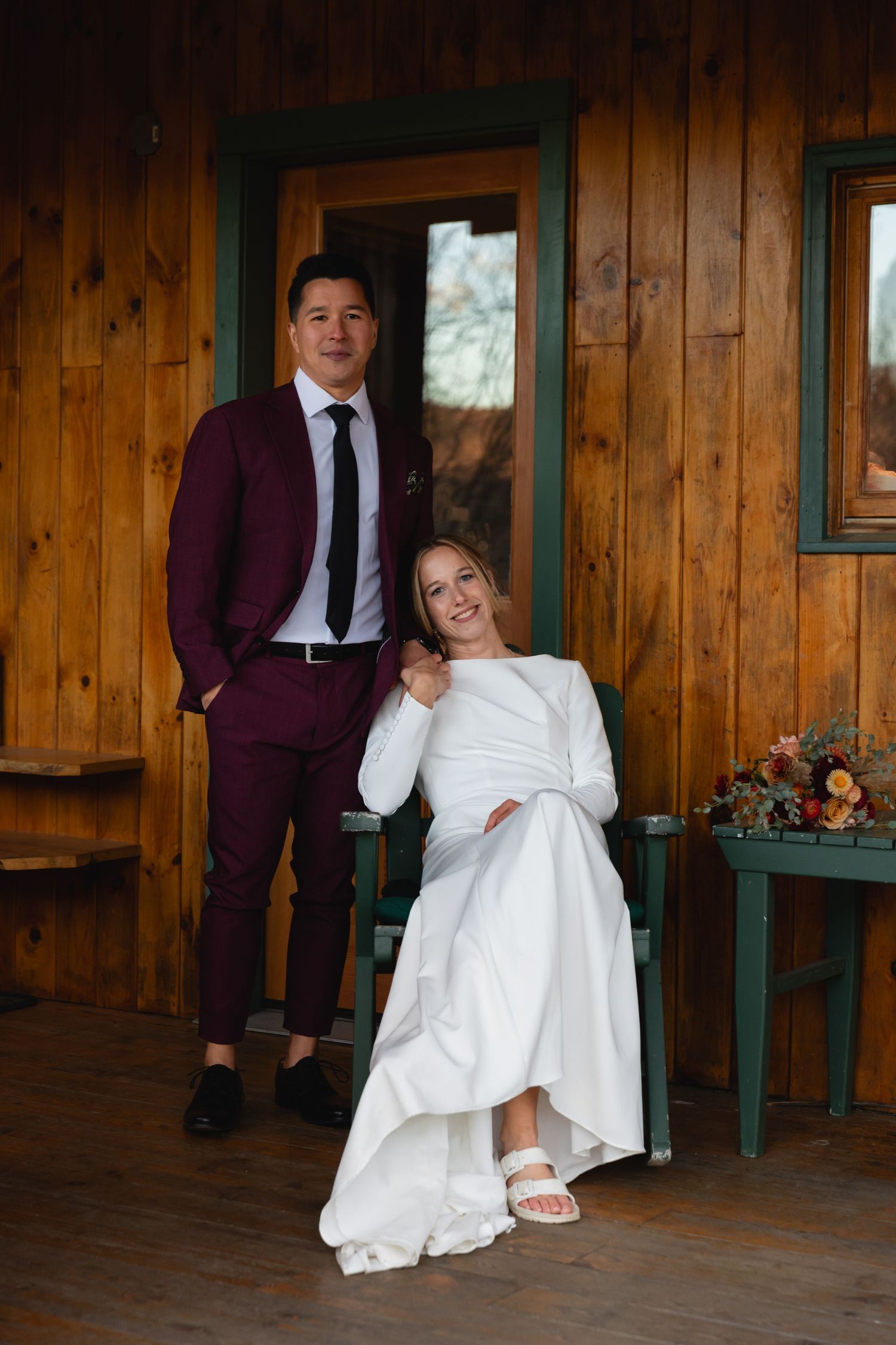 A wedding couple poses on a rustic wooden porch with burgundy suit and white dress against natural wood paneling.