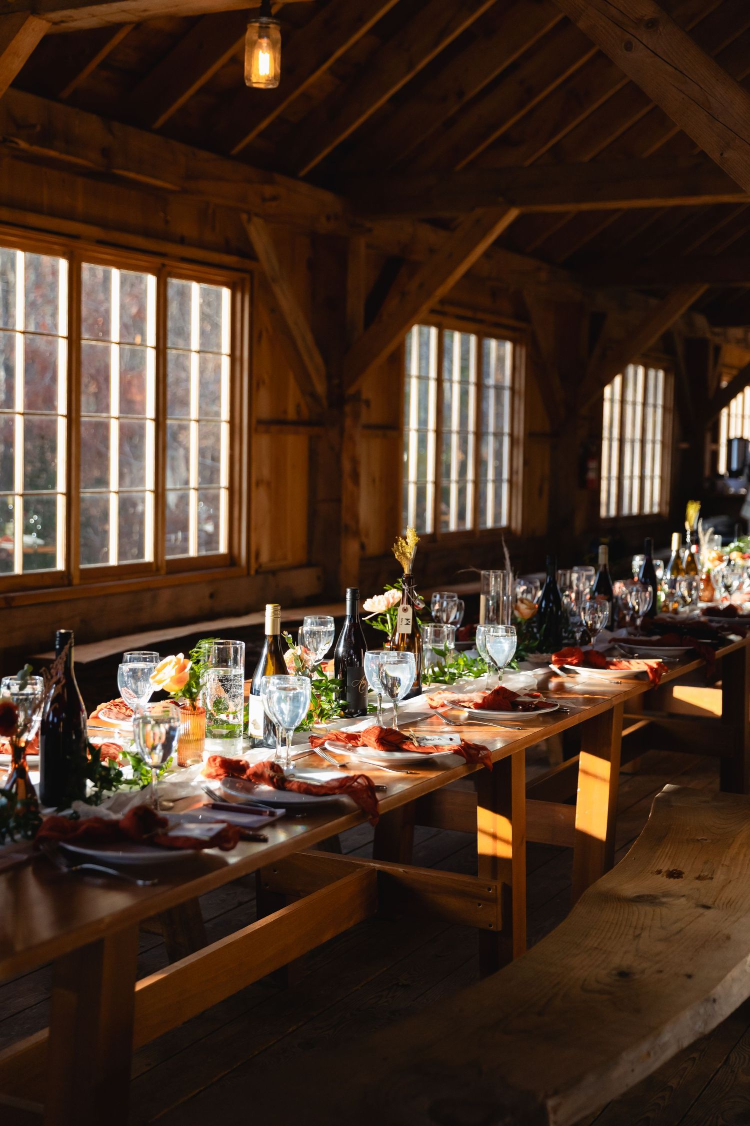 A rustic wooden barn interior with a long wooden table set for dinner featuring elegant place settings and glowing light.