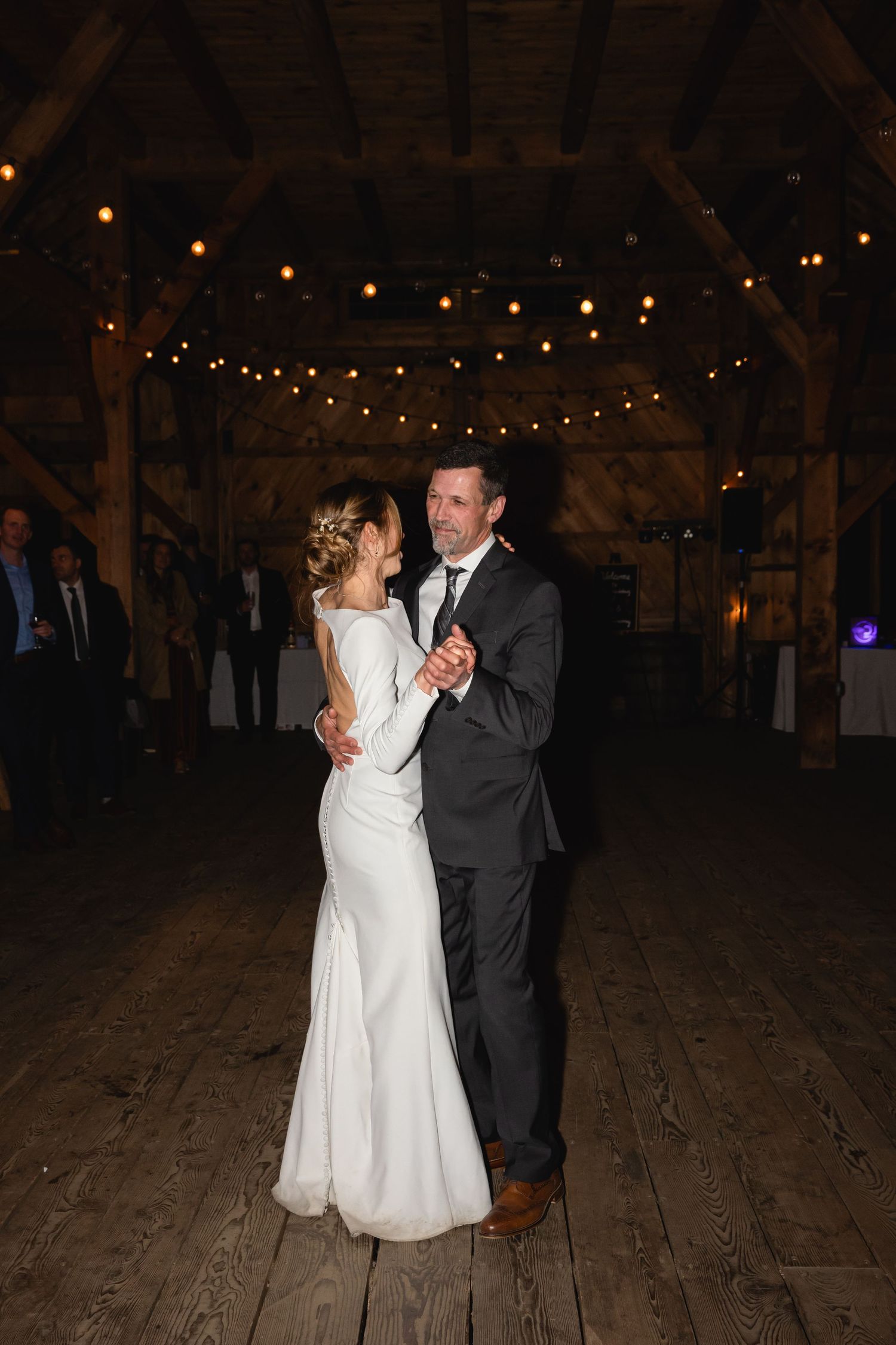 A couple shares their first dance on a rustic wooden floor under romantic string lights in a barn venue.