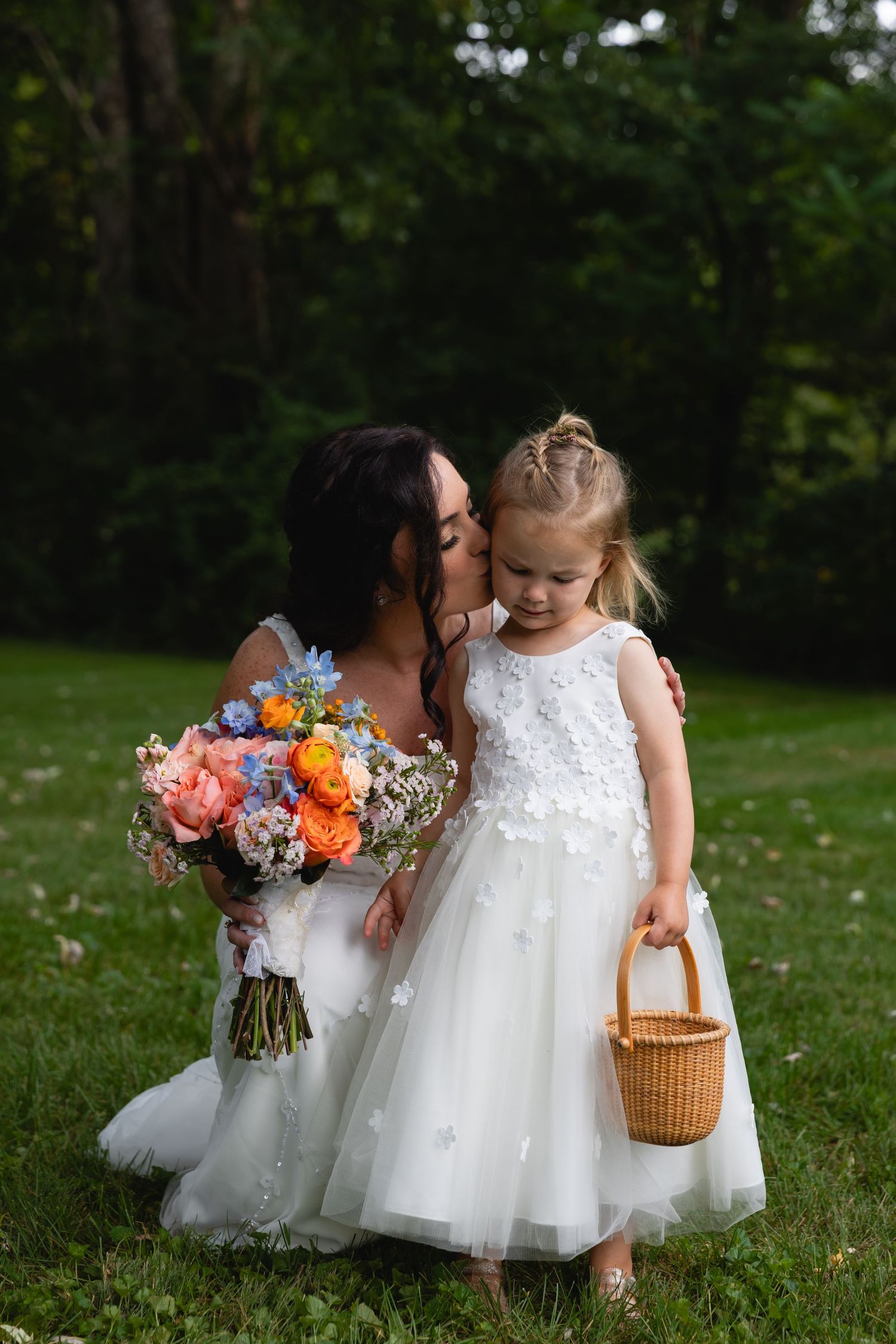 A bride and flower girl in white dresses stand together outdoors, with a colorful bouquet and wicker basket.