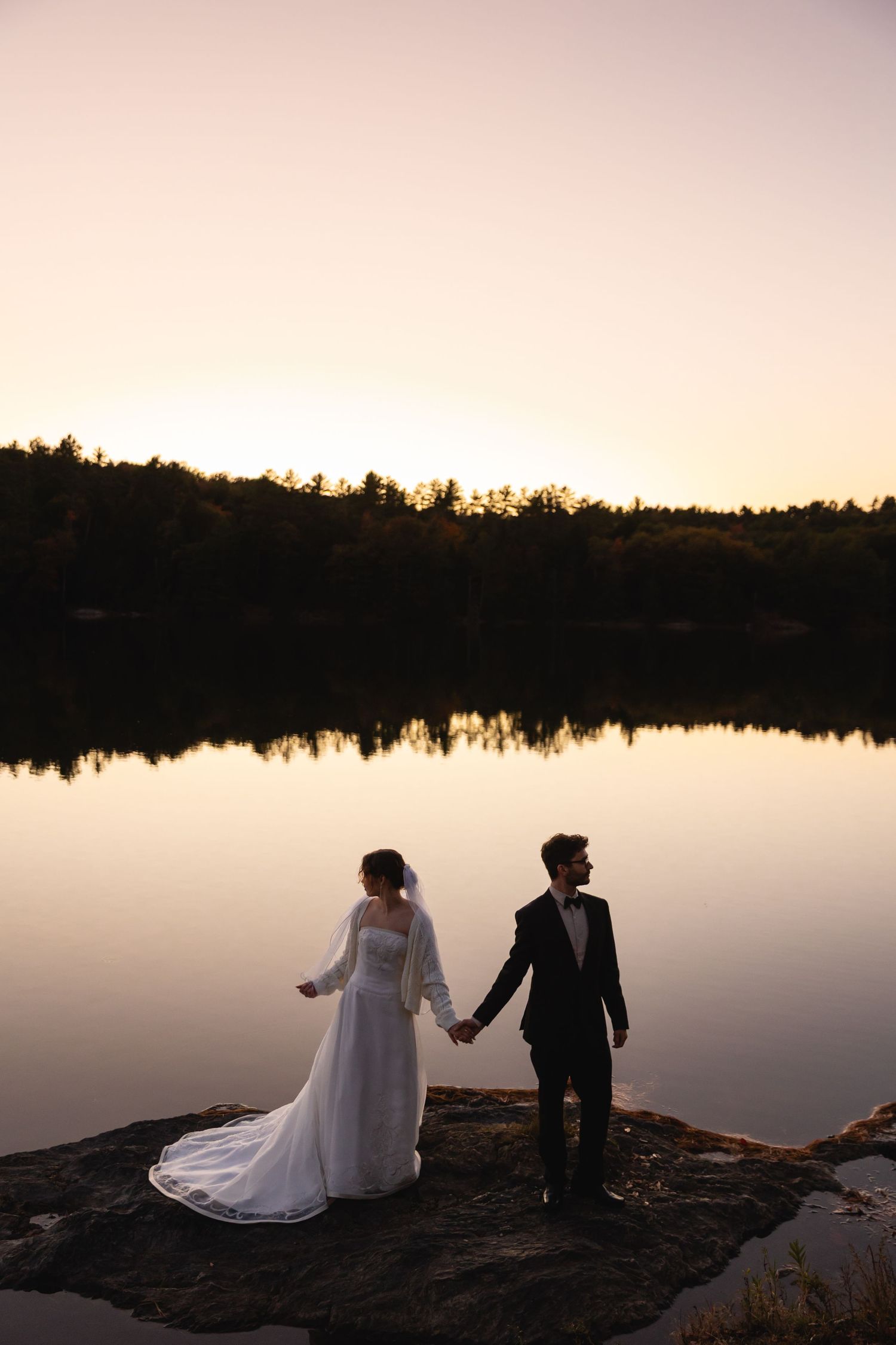 Bride and groom hold hands on rocky lakeside at sunset with mirror-like water reflection.