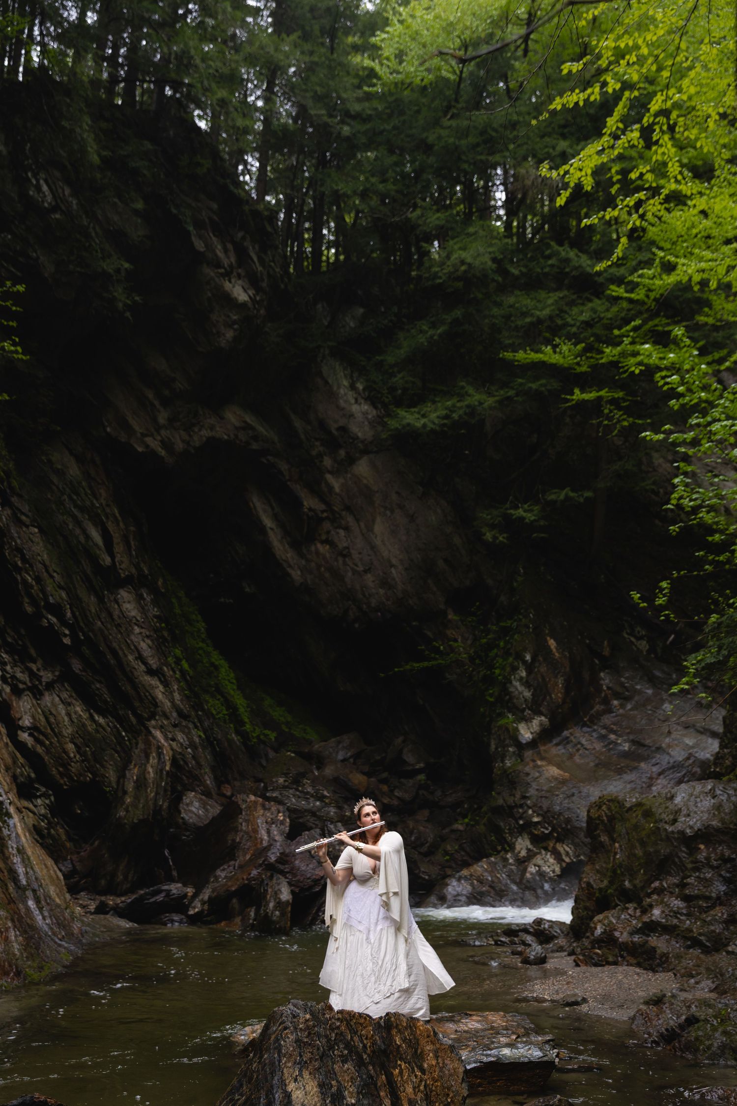 A woman in a white dress plays flute while standing on rocks beside a forest stream.