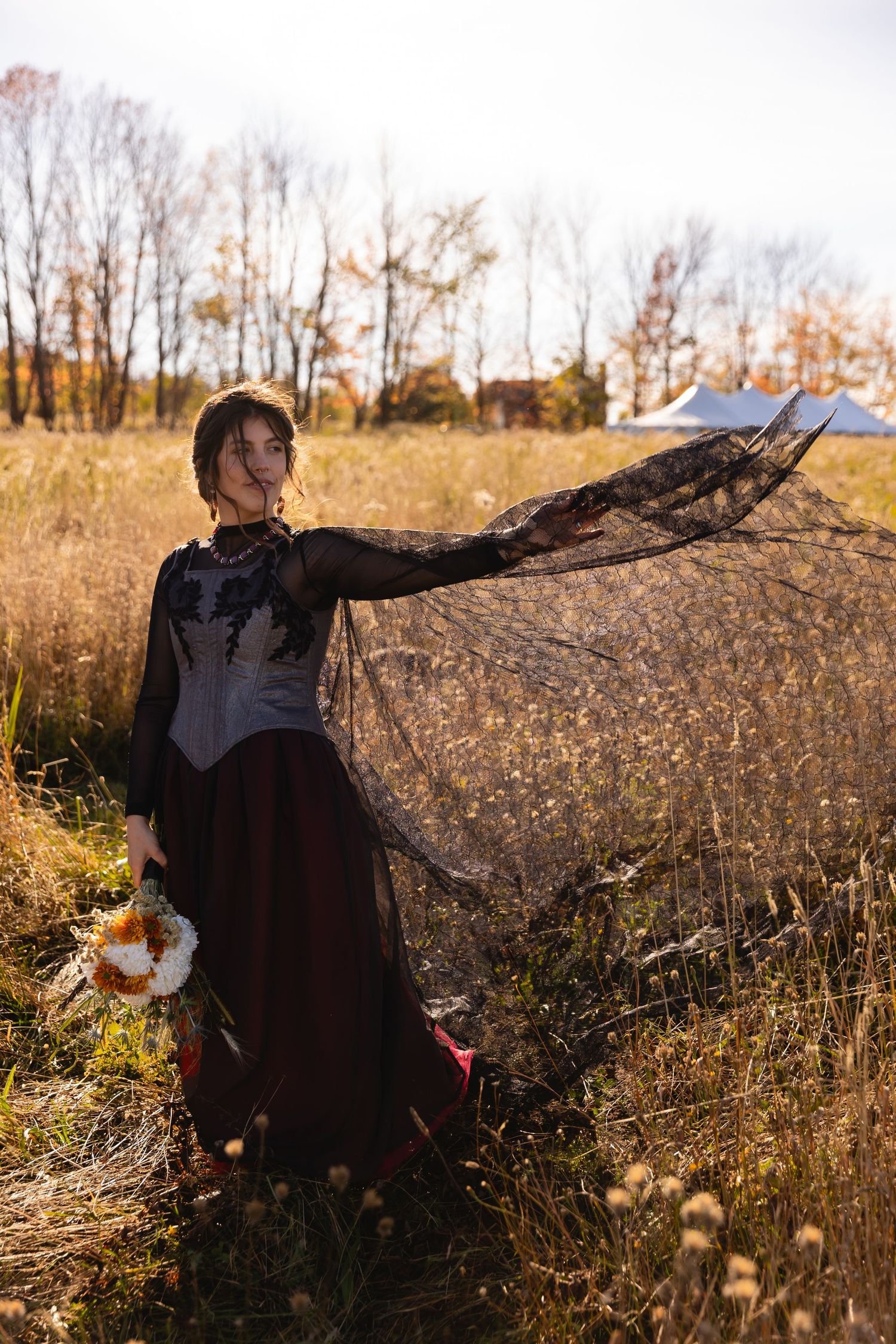 A figure in a vintage-style dress and flowing black lace veil stands gracefully in a golden autumn field at sunset.
