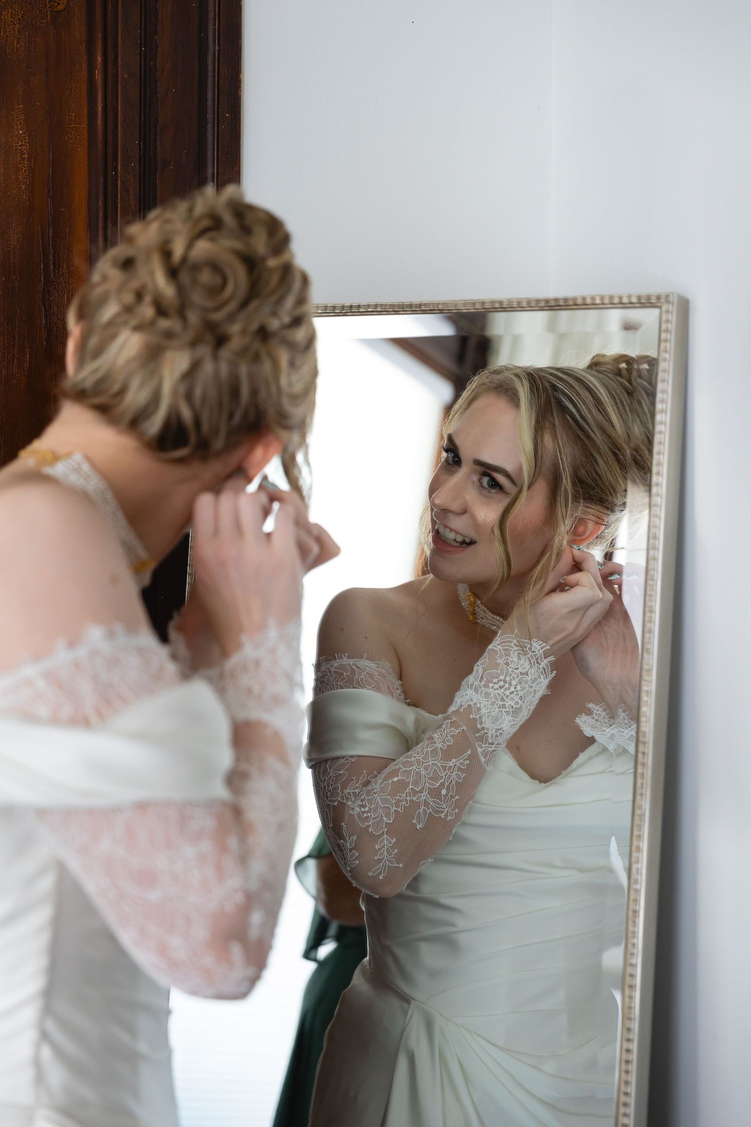 A bride in an elegant white gown and lace sleeves looks at her reflection in an ornate framed mirror.