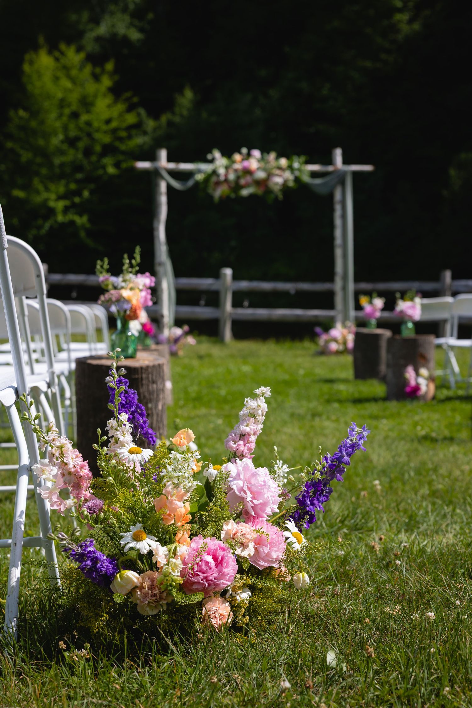 Colorful floral arrangements line a grassy outdoor aisle with white chairs and fencing for a garden wedding ceremony.