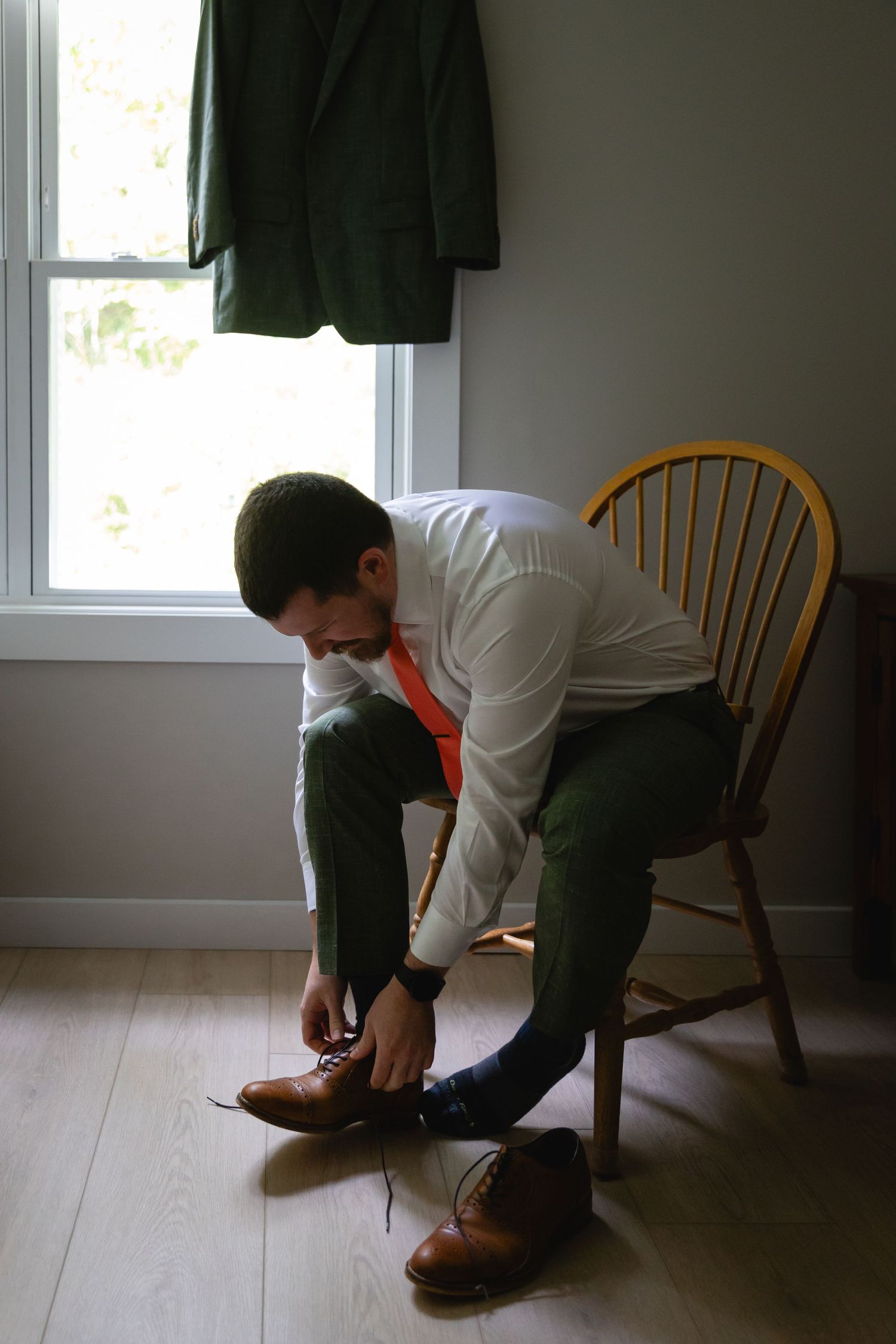 Someone tying brown leather dress shoes while sitting on a wooden chair near a window.