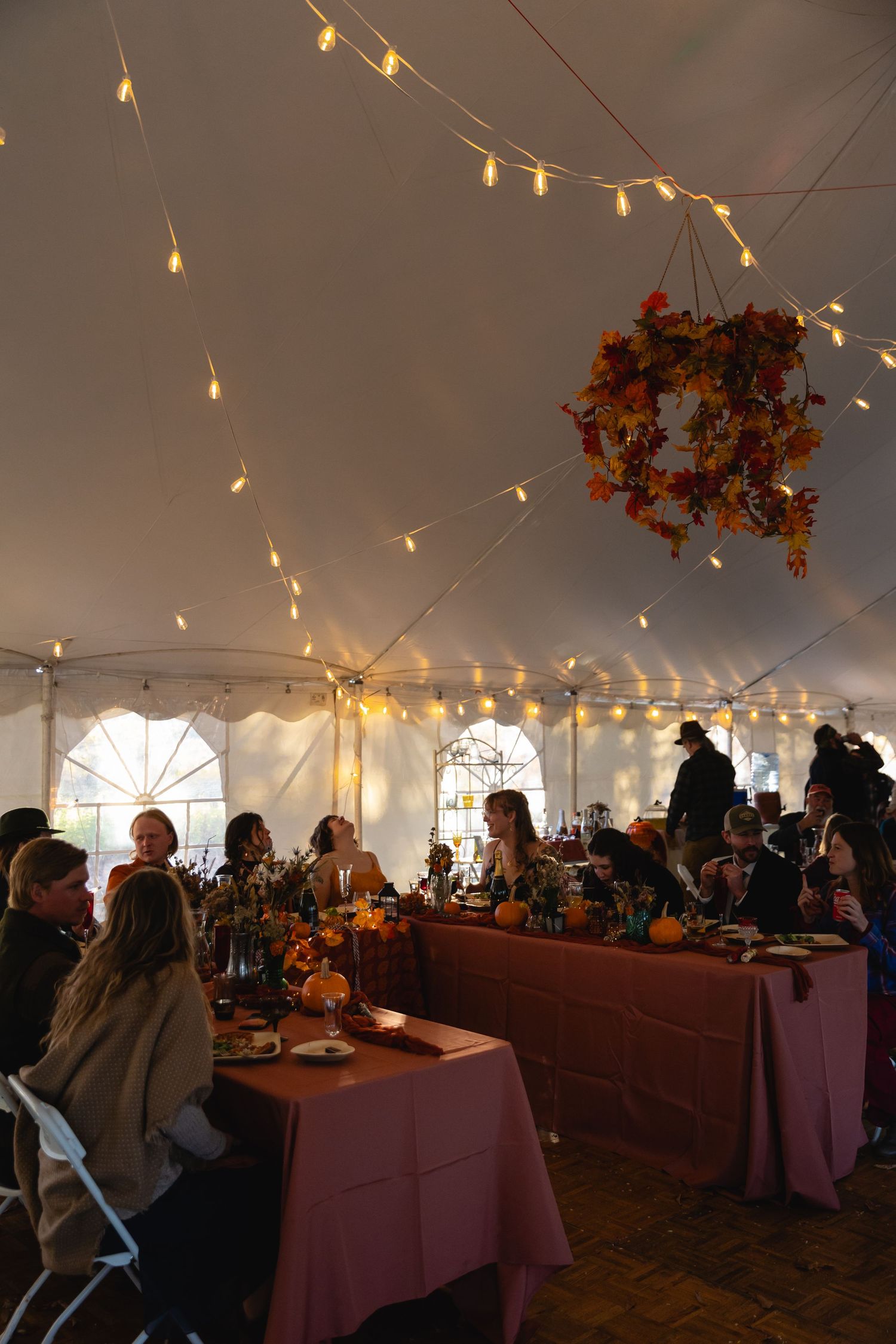 Wedding reception setup under white tent with string lights and burgundy tablecloths creating an elegant autumn atmosphere.