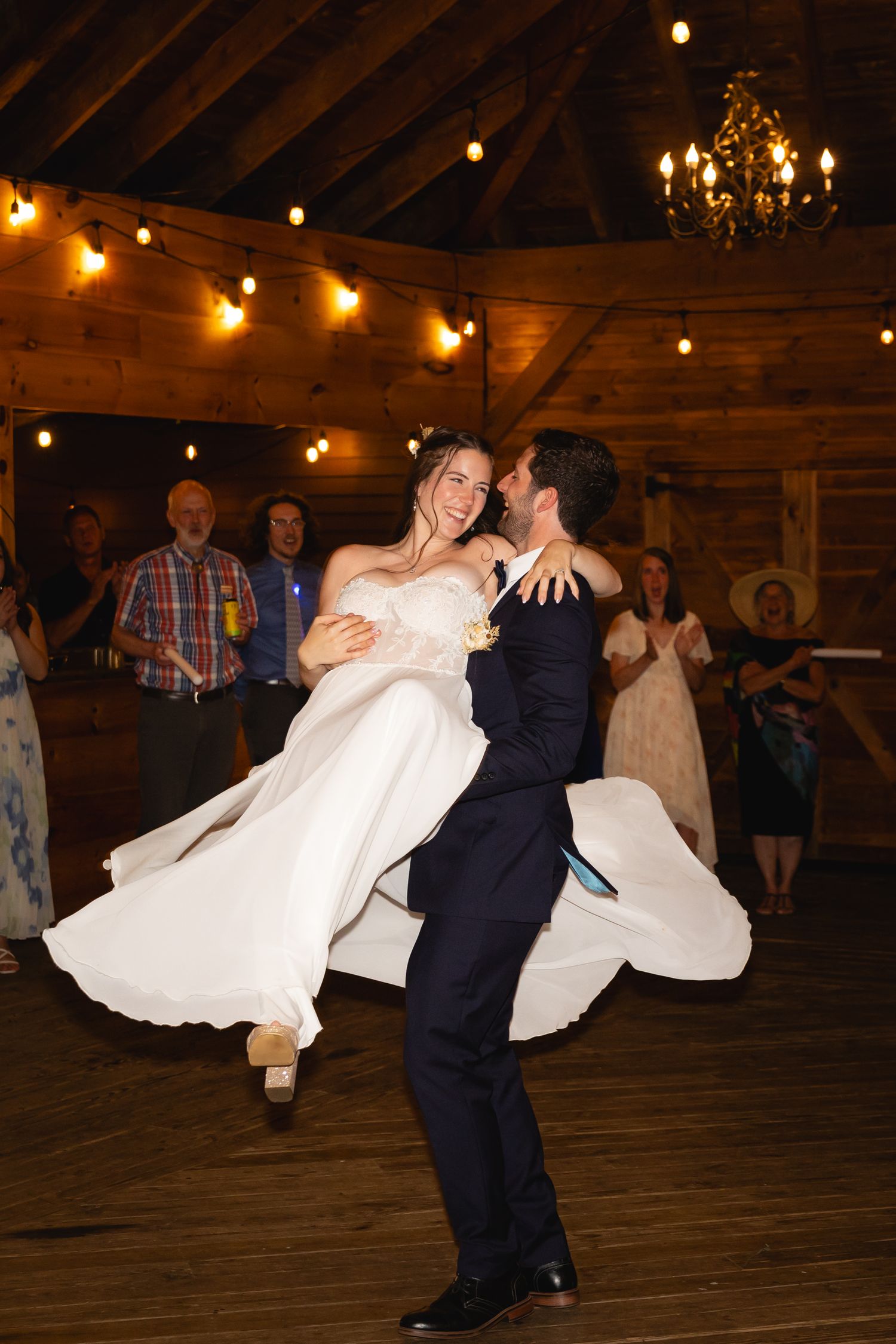 A couple dances their first dance in a rustic barn venue with string lights and a chandelier overhead.