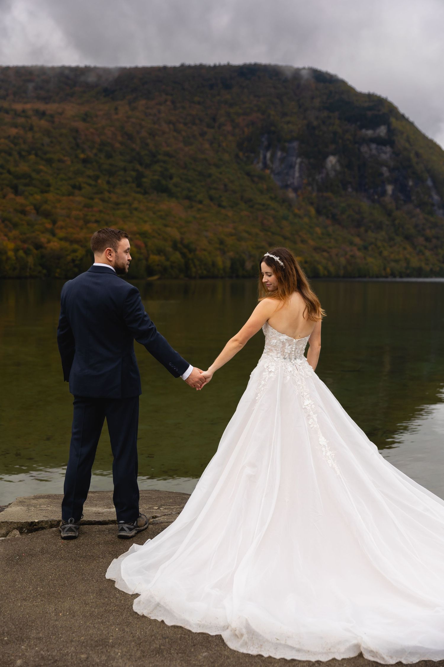 Bride in flowing white gown holds groom's hand by mountain lake with fall foliage.