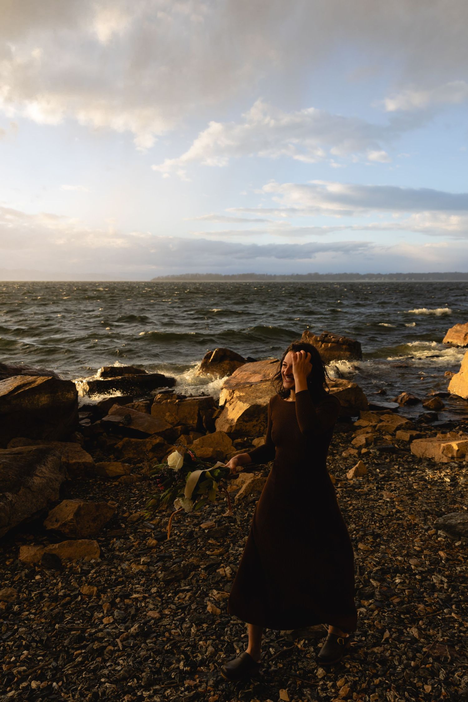 Person in a dark dress stands on rocky coastline at dusk with dramatic clouds and ocean waves.