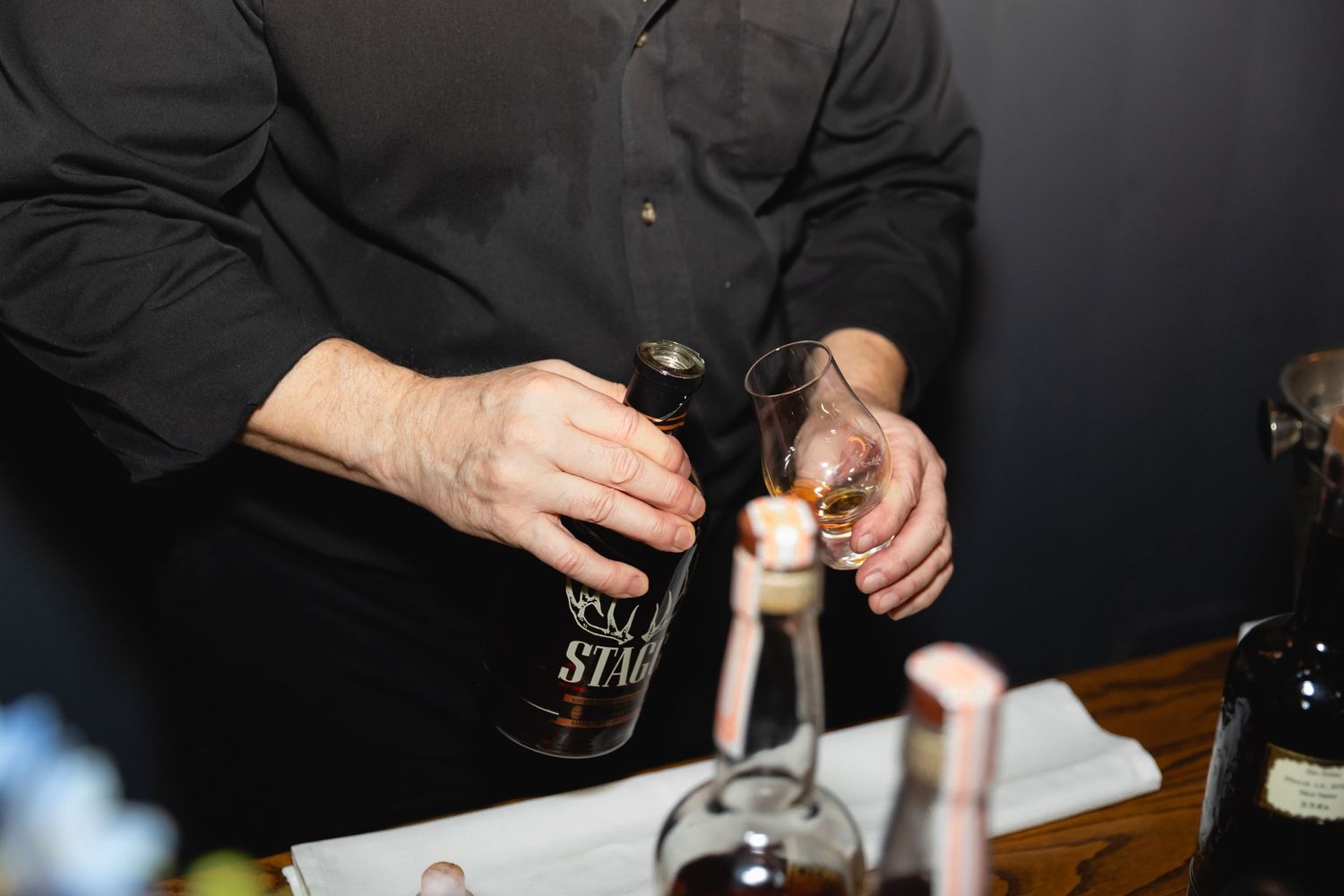 Close-up shot of hands pouring Stella Artois beer from bottle into glass at dark table setting.