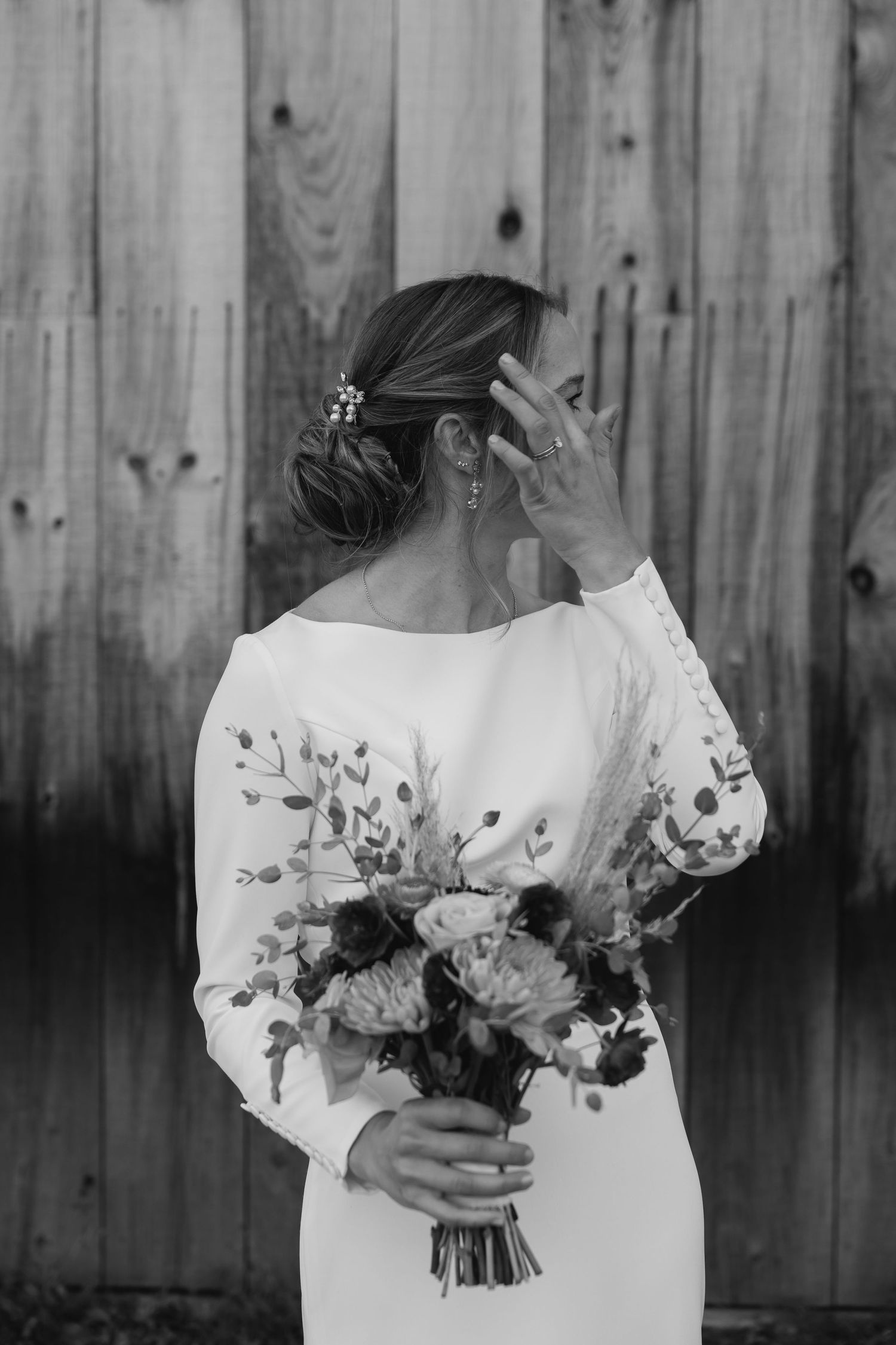 Black and white photo of bride in long sleeve dress holding floral bouquet against wooden fence background.