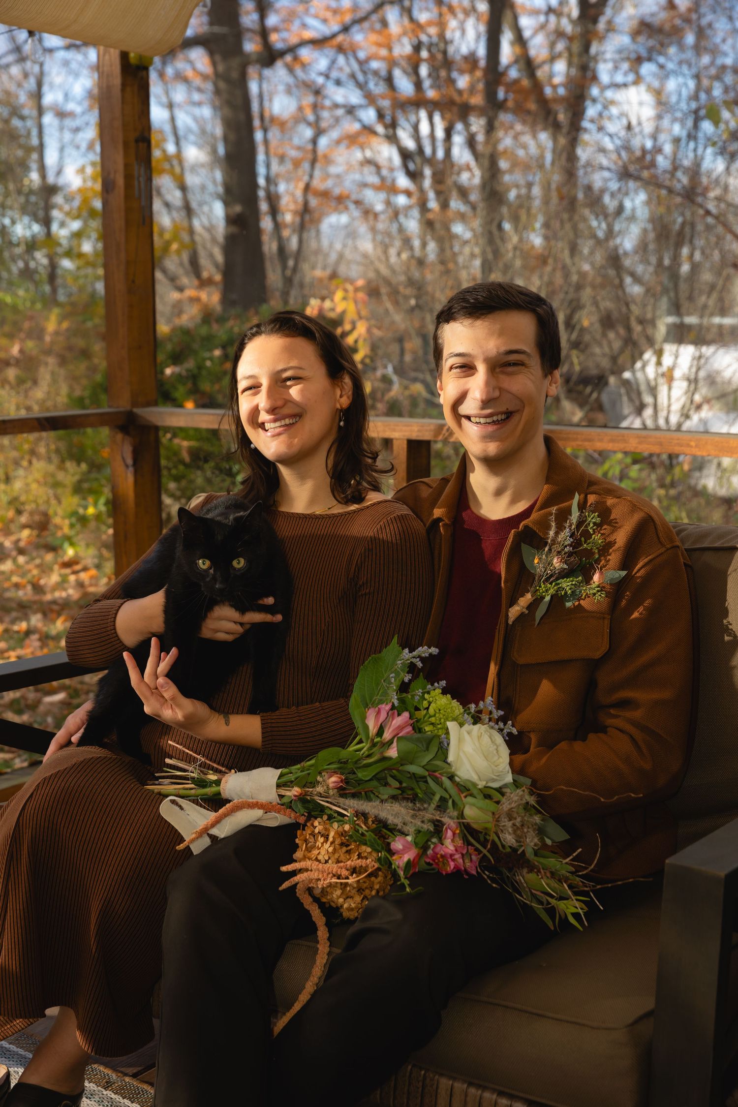 A couple sits on a porch bench holding a black cat and floral bouquet in autumn woodland setting.