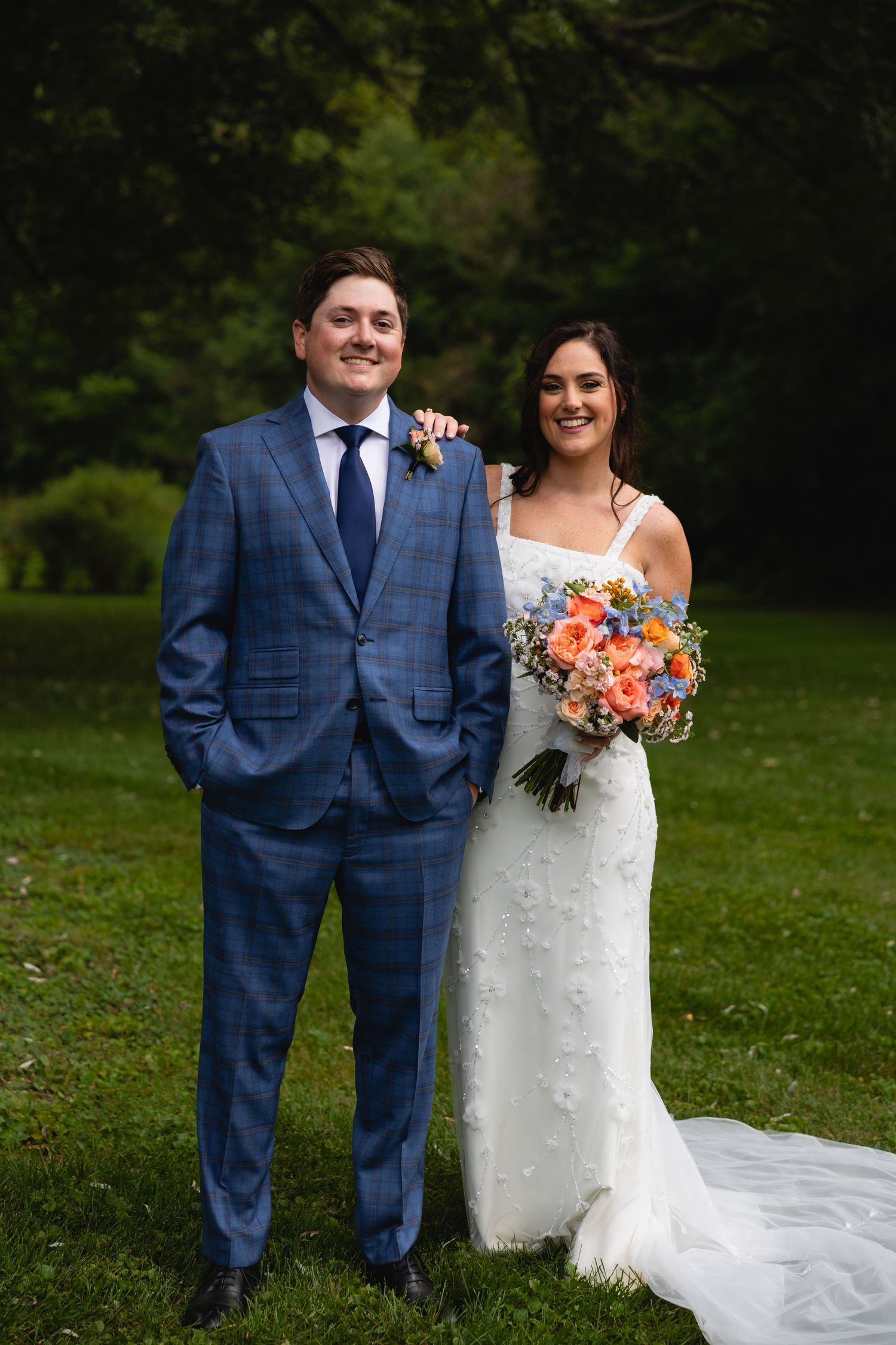 A wedding couple poses together outdoors, with the bride holding a vibrant coral and peach bouquet.