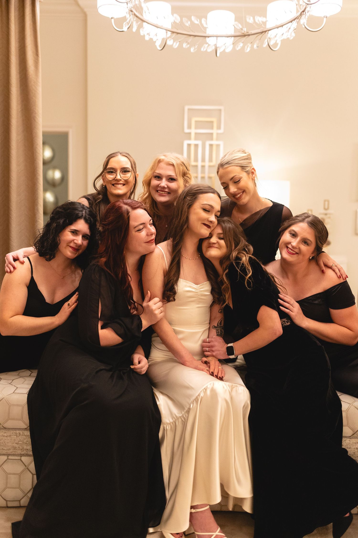 Bridesmaids in black dresses gather around bride in white gown for candid wedding photo under warm lighting.