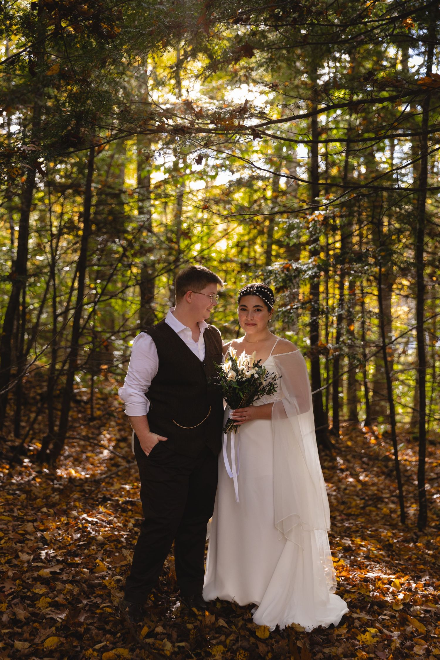 Wedding couple poses in formal attire against a backdrop of sunlit autumn forest trees.