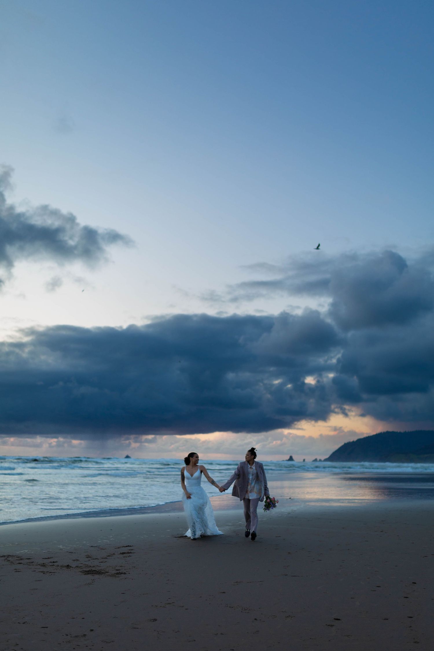 A couple walks hand in hand along a beach at sunset with dramatic clouds overhead.