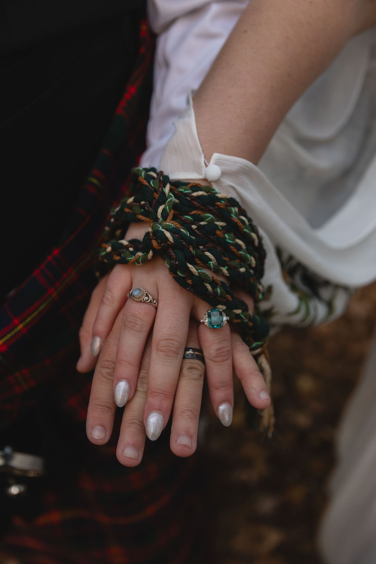 Closeup of hands with rings bound by a Celtic handfasting cord against tartan fabric.
