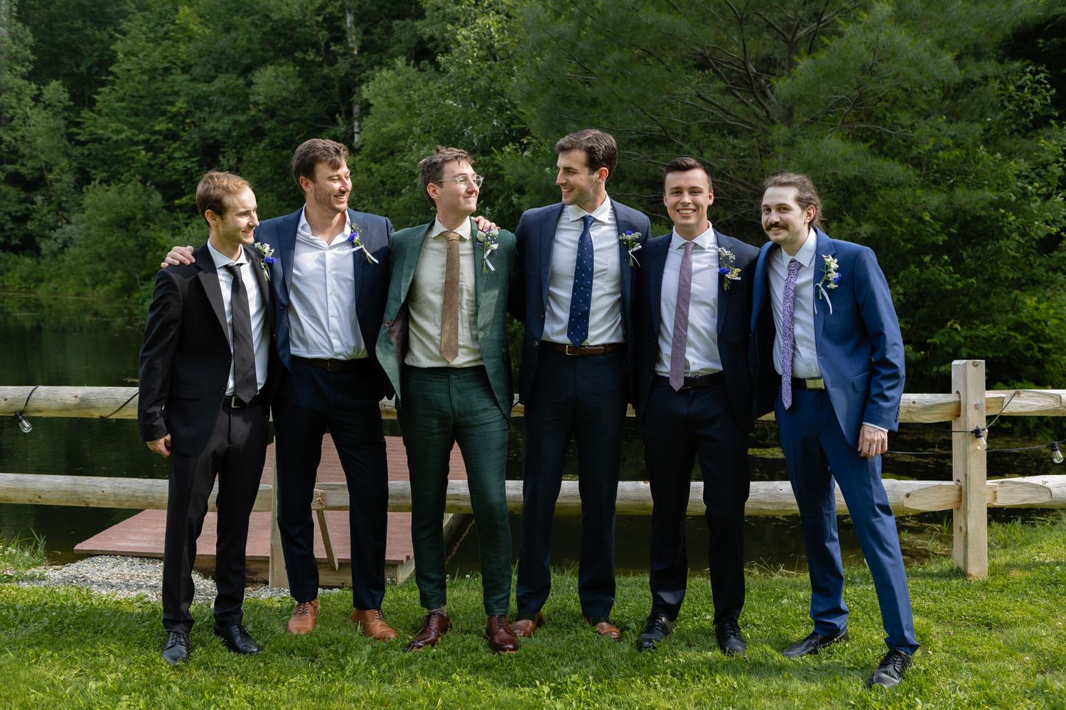 Group of groomsmen in mixed formal attire share laughs while posing outdoors.
