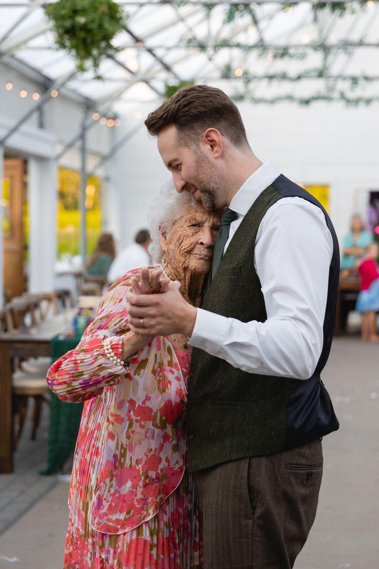 Groom tenderly dancing with elderly grandmother at elegant greenhouse wedding reception.