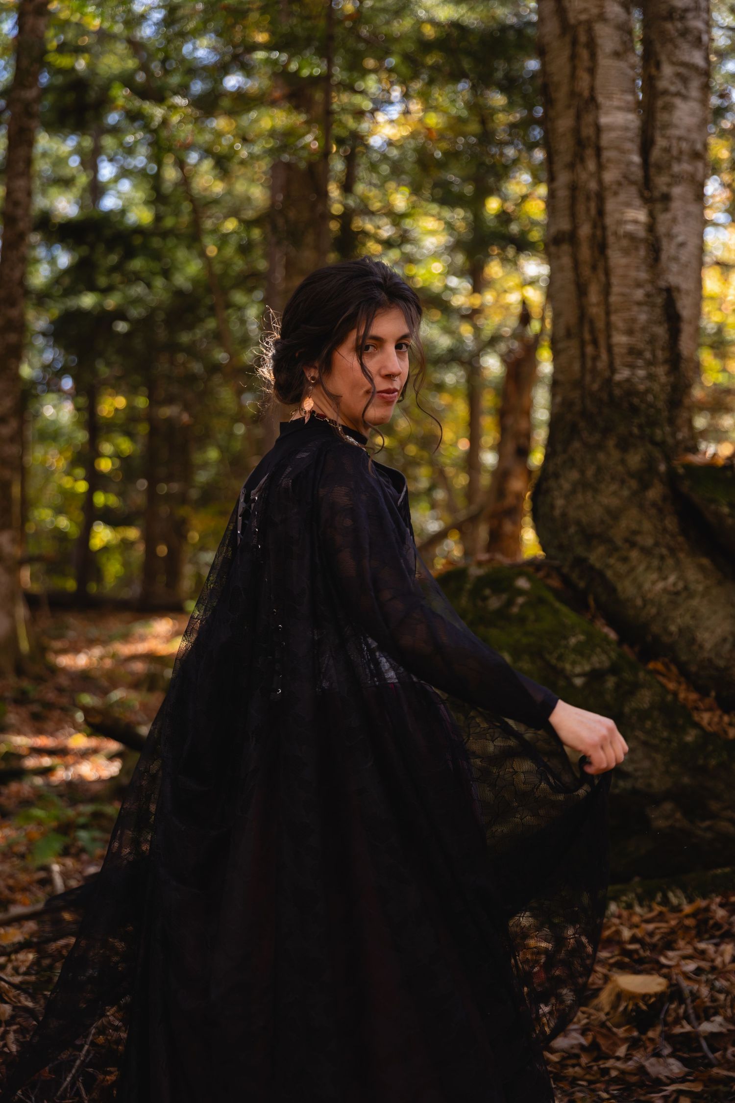 A figure in a flowing black dress twirls dramatically through an autumn forest setting with fallen leaves on the ground.