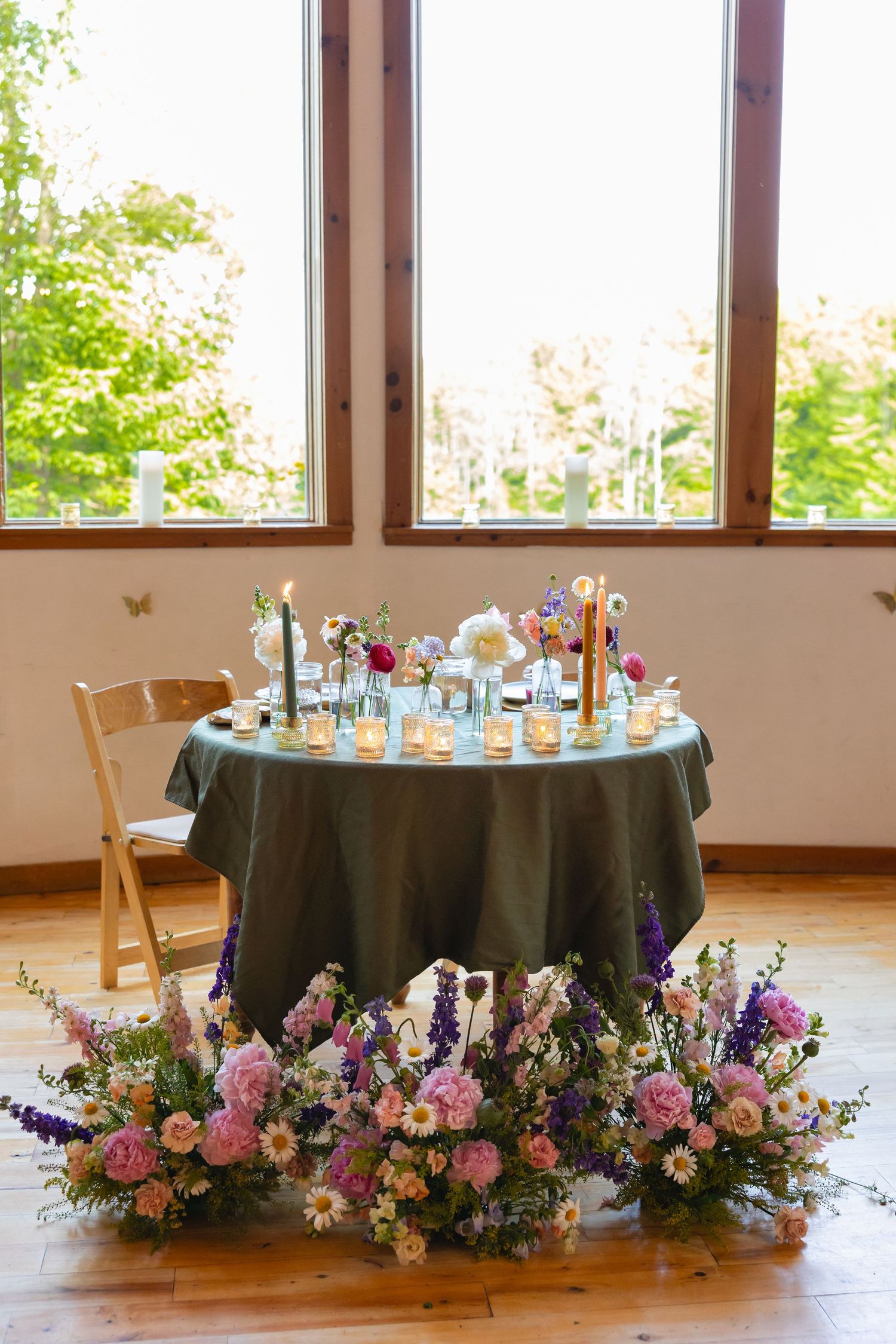 Round tables with dark green tablecloths decorated with pink and purple floral arrangements in a sunlit venue.