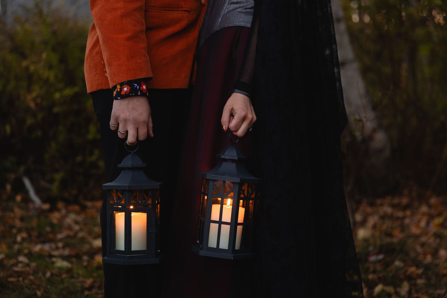 Two glowing lanterns held in hands against a dark autumn background with fallen leaves on the ground.
