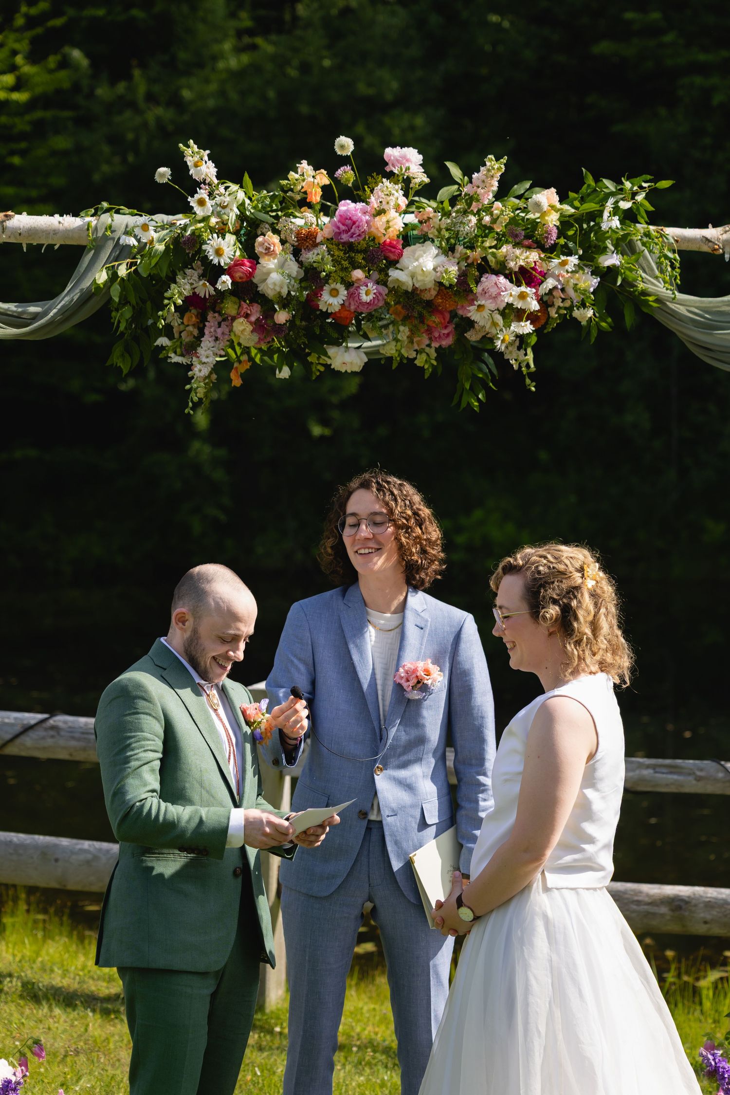 Outdoor wedding ceremony setup with floral arch decorations and guests celebrating in a sunny garden setting.