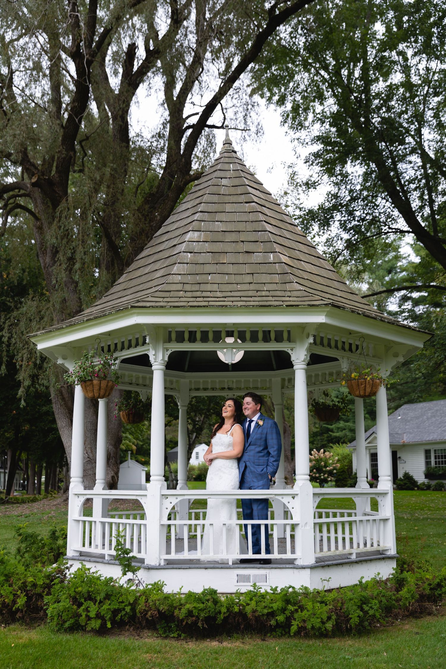 A classic white wooden gazebo with ornate trim and shingled roof stands among trees and hanging moss in a garden setting.