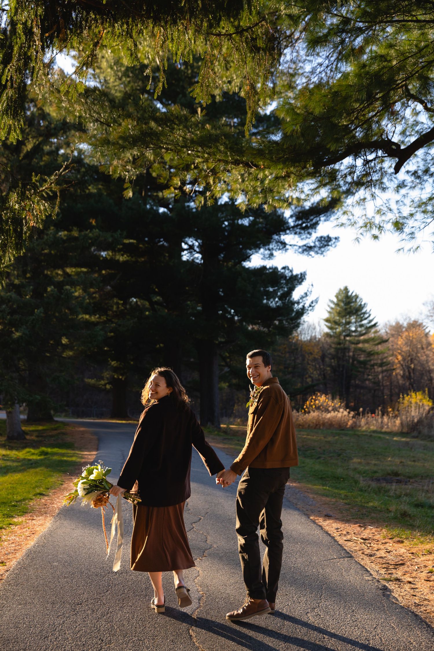 A couple walks together on a sunlit path through evergreen trees on an autumn afternoon.