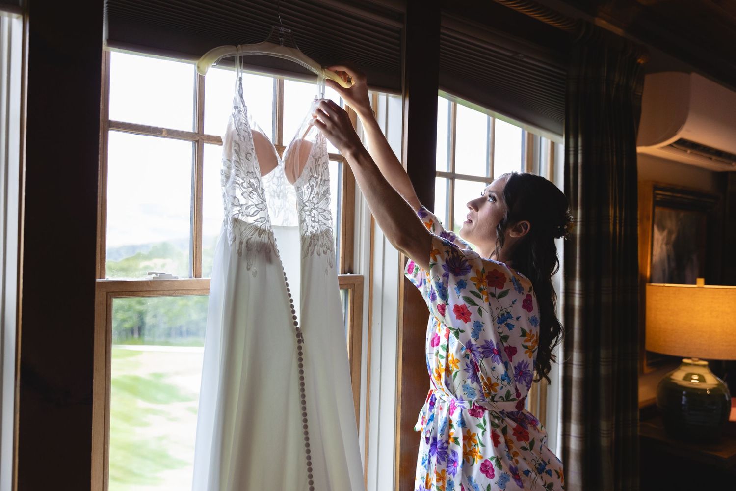 White lace wedding dress being held up against sunlit windows.
