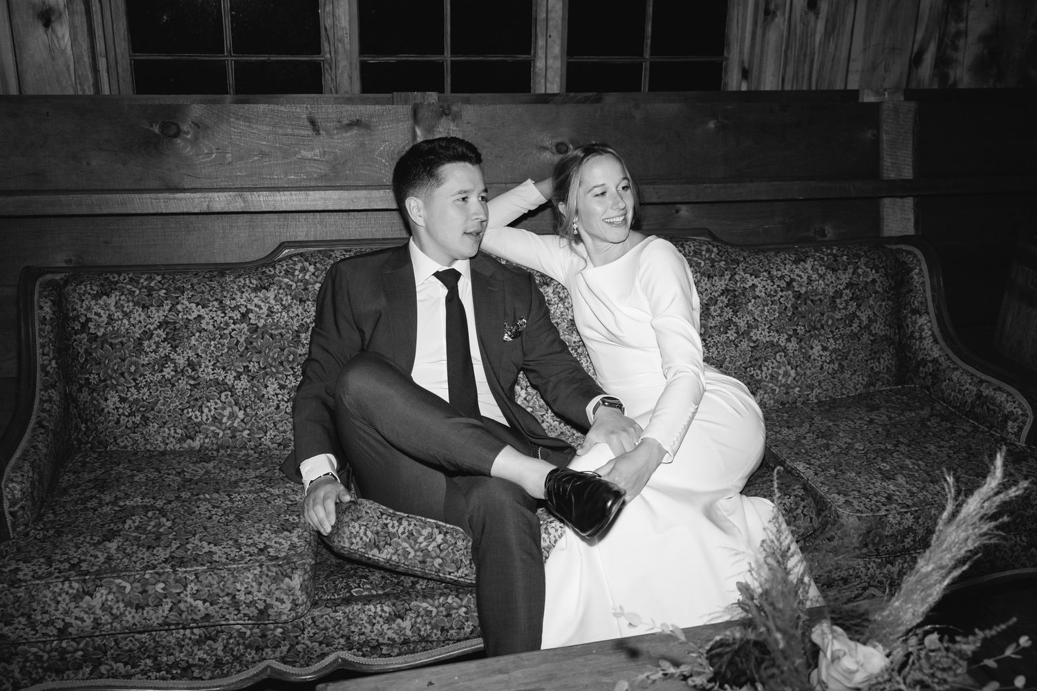 A couple in formal wedding attire sits together on a vintage floral couch in a black and white photograph.