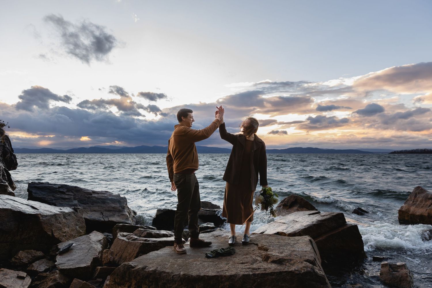 Two people high-five while standing on rocky coastal outcrop at sunset with dramatic cloudy sky and ocean waves.