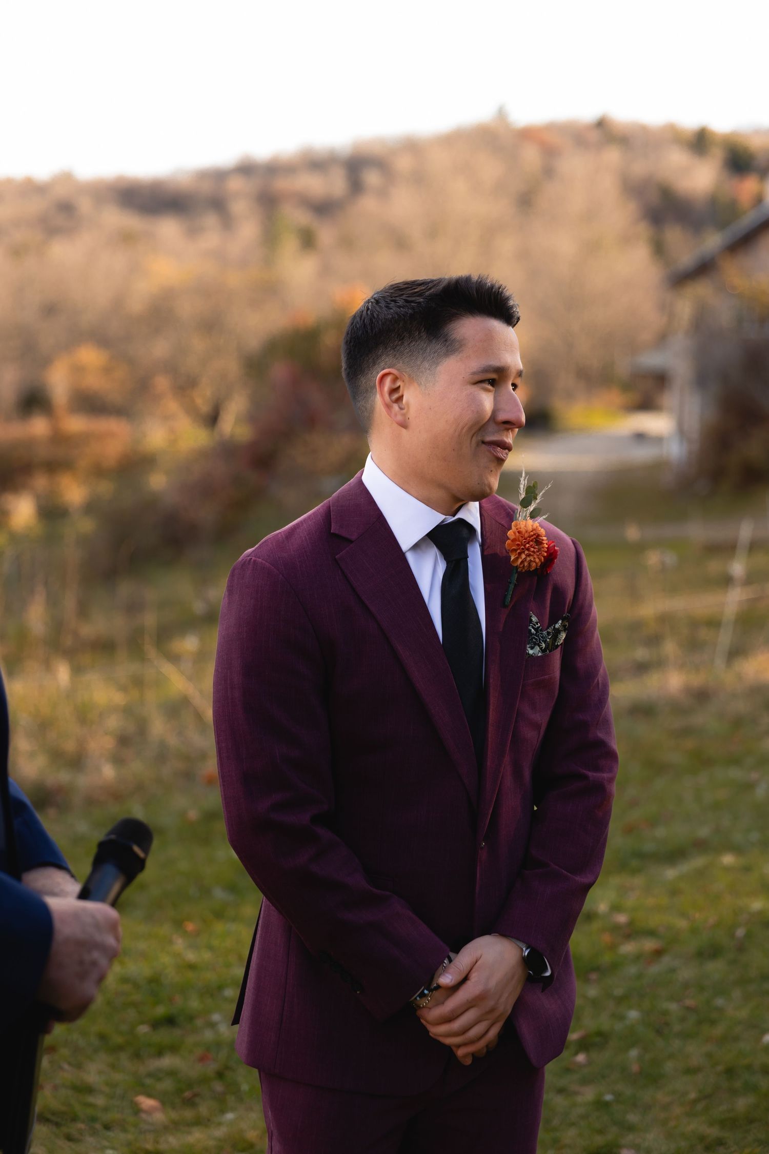 A wedding attendee in a burgundy suit and navy tie smiles during an outdoor autumn ceremony.