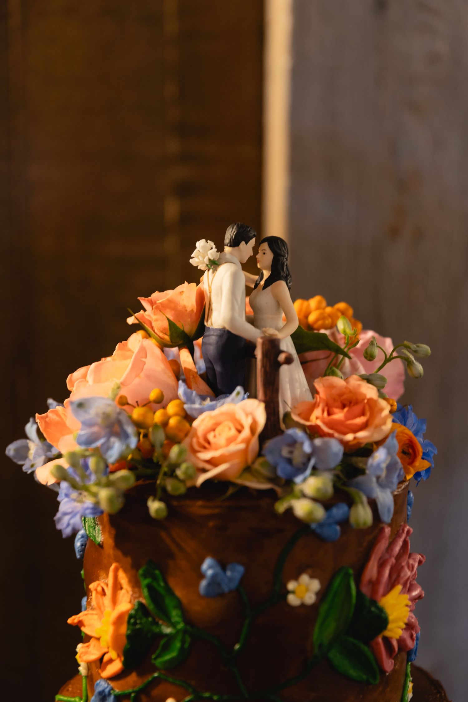 Wedding cake topper features a romantic couple surrounded by peach roses and blue delphinium flowers.