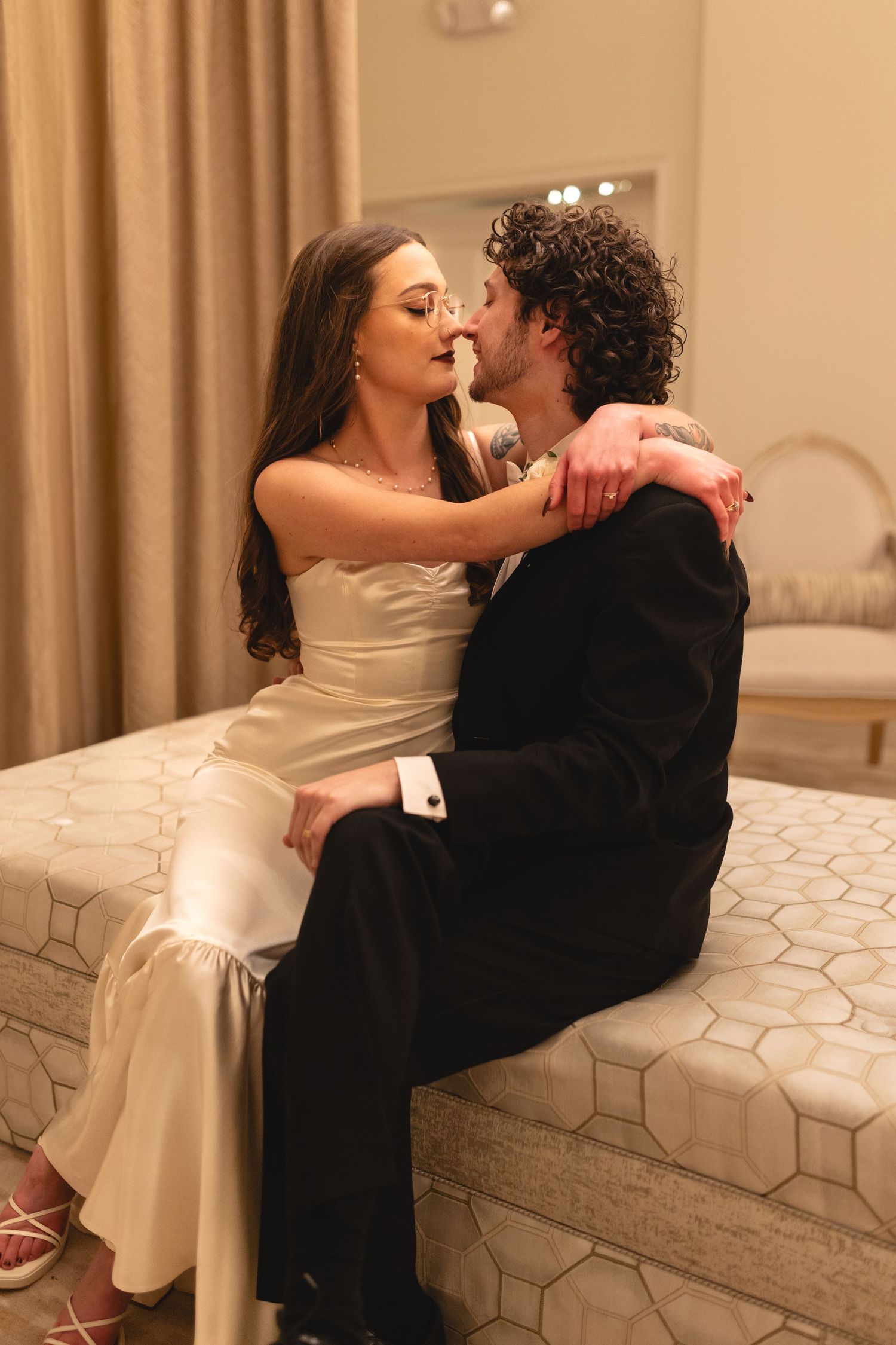 A romantic couple shares an intimate moment while sitting on a beige ottoman in formal evening attire.