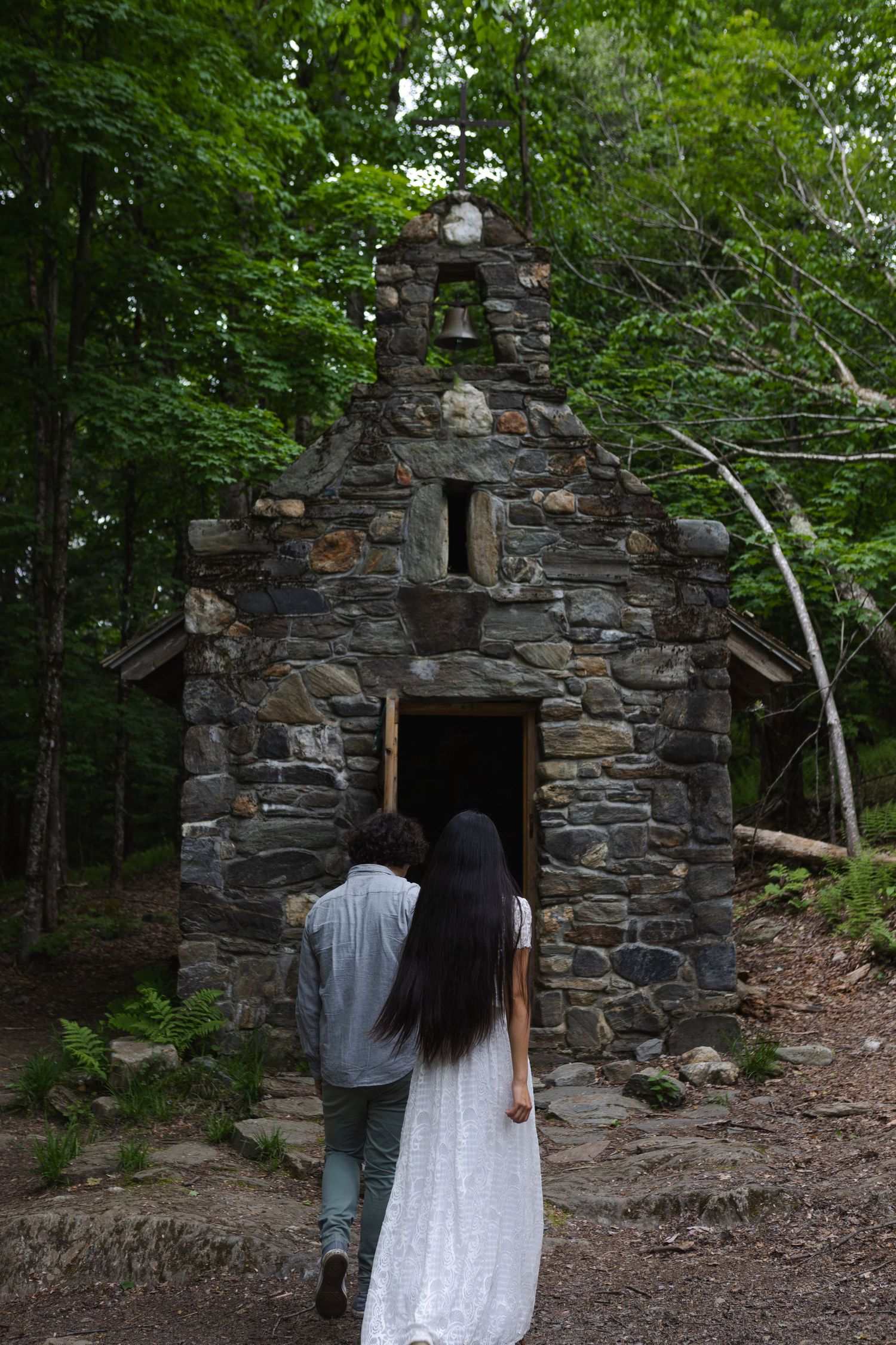 A couple approaches a rustic stone chapel entrance surrounded by dense forest.