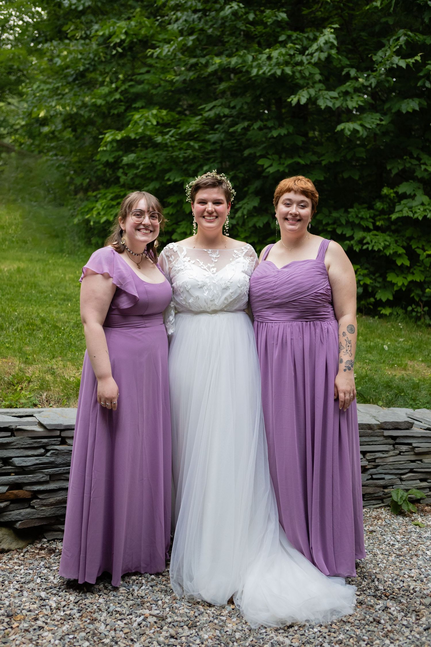 Wedding party wearing white and purple dresses pose by stone wall in garden setting.