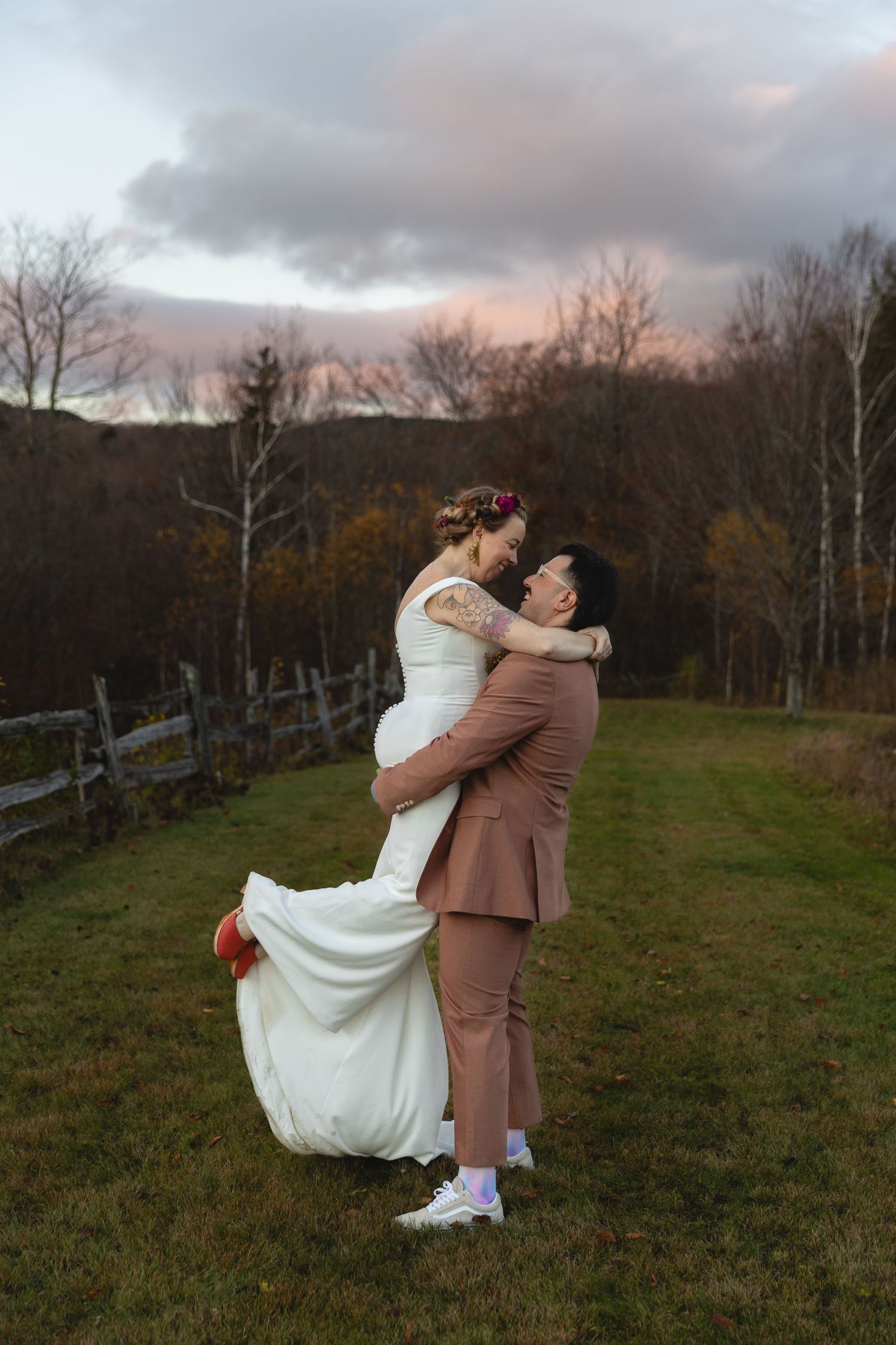 A couple shares a joyful embrace in a field during sunset with fall foliage in the background.