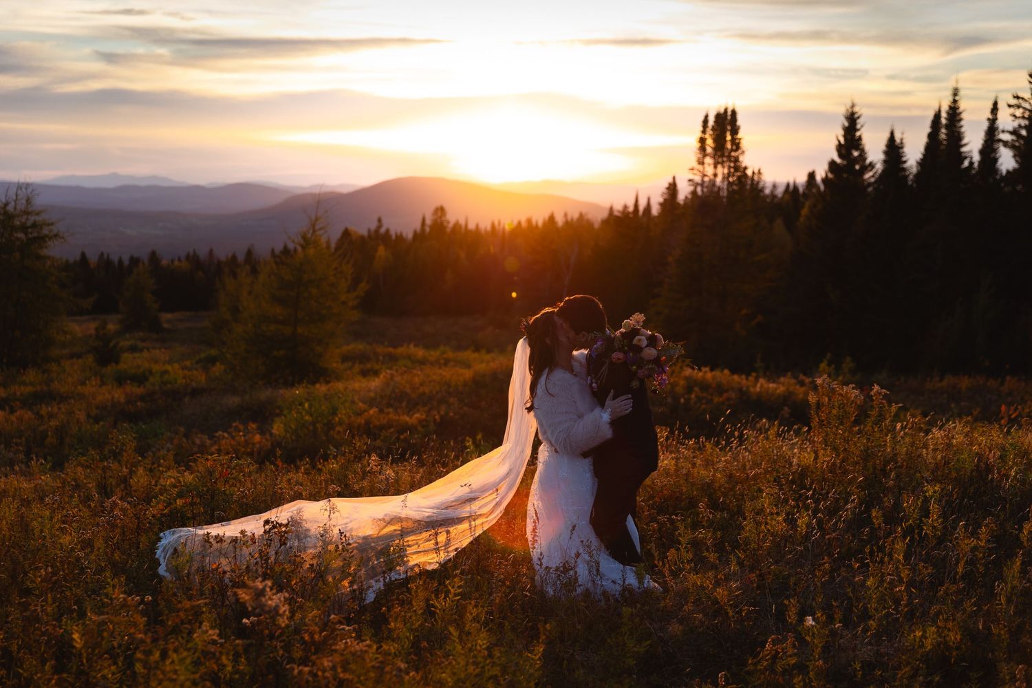 Bride and groom kiss at sunset in a wildflower meadow with mountain views and golden light.